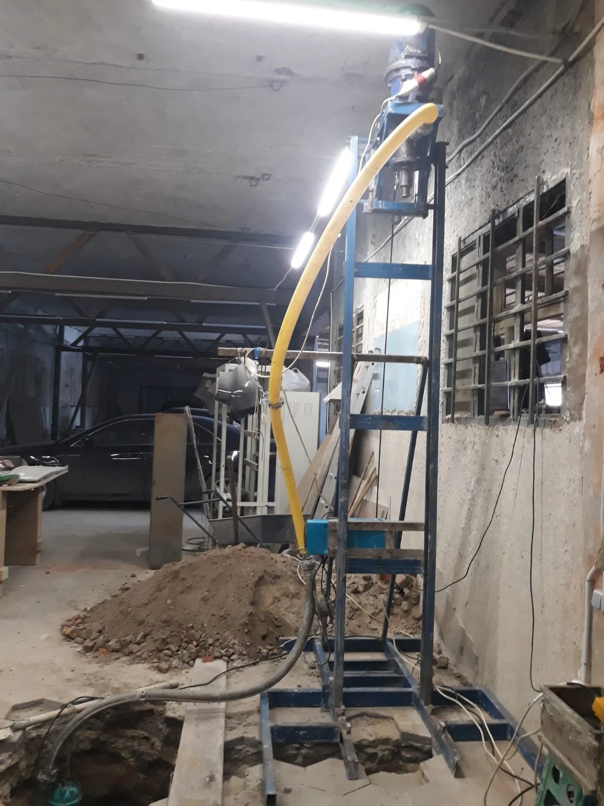 Interior of a construction site with exposed concrete walls and ceiling, a black car parked on the left side, a pile of dirt on the floor in front, and construction equipment and tools scattered around, including a blue scaffold and a yellow hose.