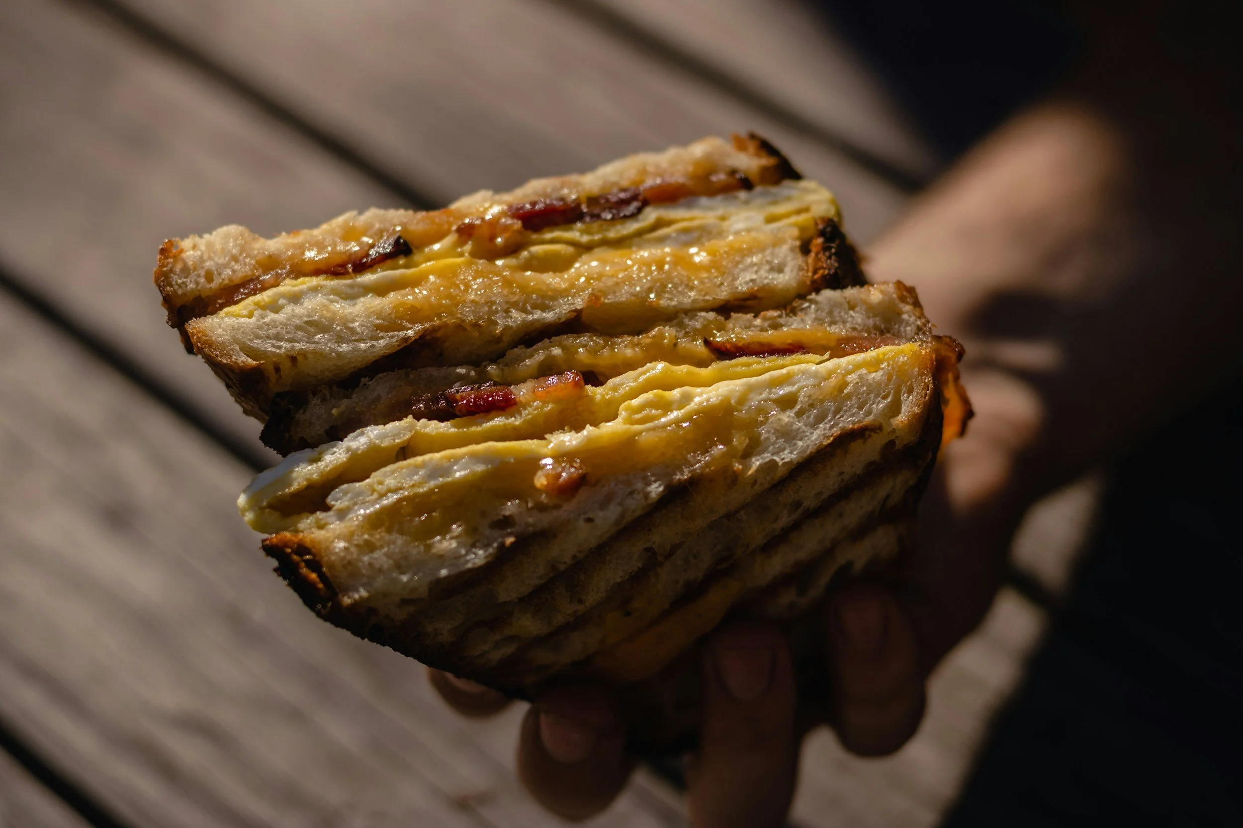 Close-up of a sandwich with melted cheese, bacon, and toasted bread, being held over a wooden surface.