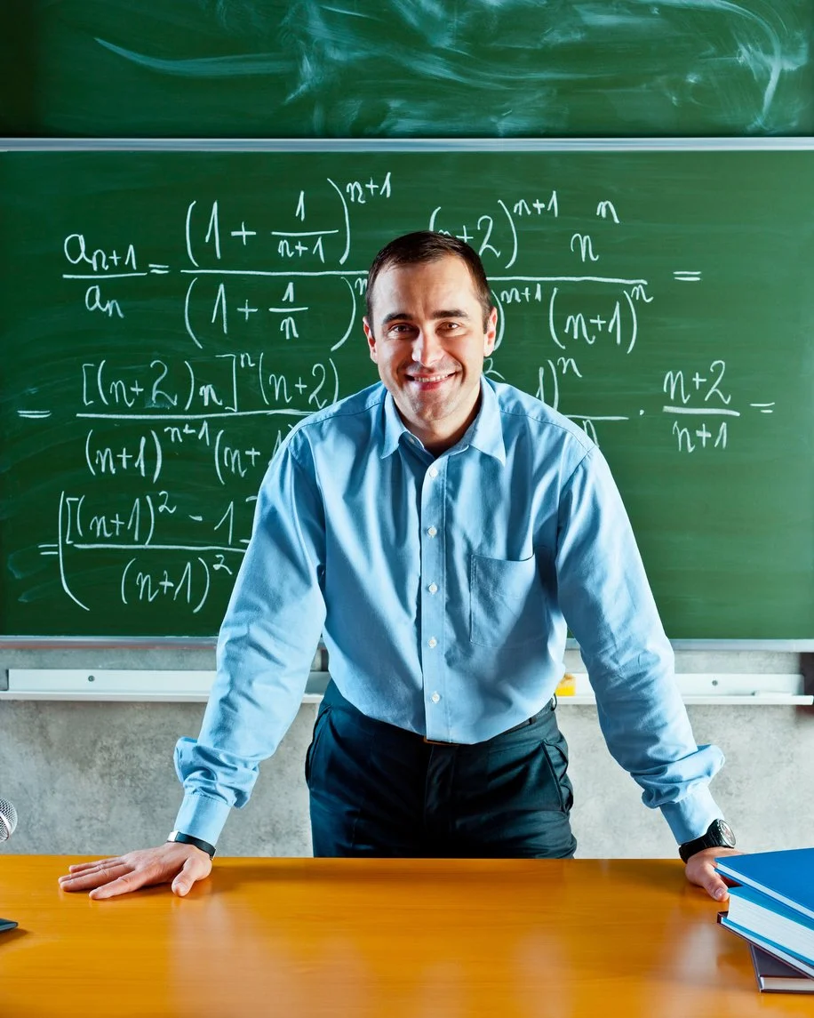 A male school teacher smiling in front of a blackboard covered in math equations, representing professional environments with restricted bathroom access.