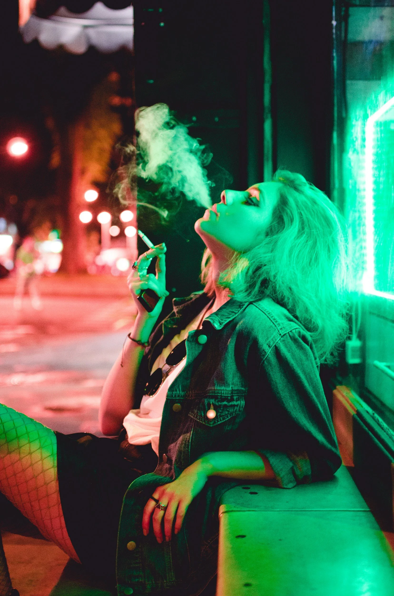A woman smoking a cigarette under a green neon light, illustrating environmental tobacco smoke exposure as a vital factor in an individual's health portfolio.