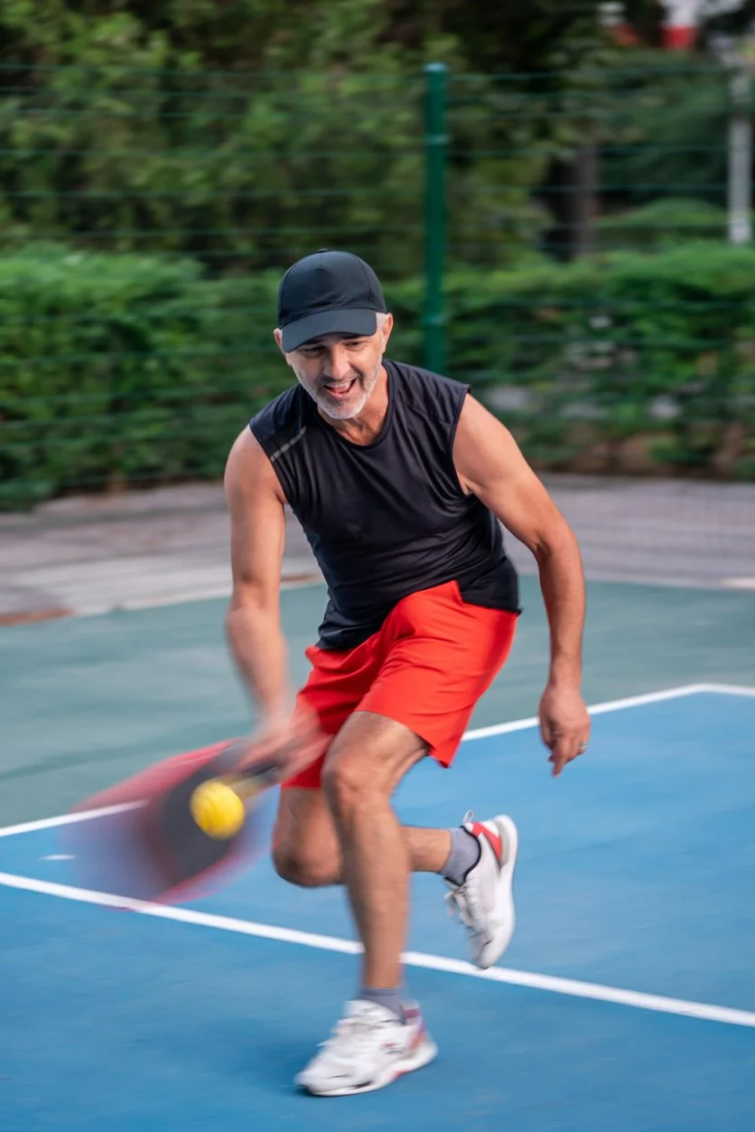 An active middle-aged man in red shorts playing pickleball on an outdoor court, demonstrating vitality, physical health curiosity, and full-body wellness at any age.