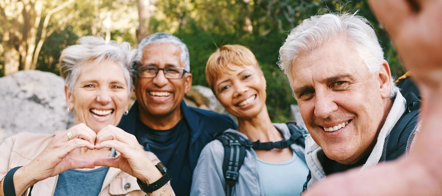 A smiling group of active seniors and middle-aged friends outdoors taking selfies with heart hands, shattering aging stereotypes through lifelong health curiosity and shared vitality