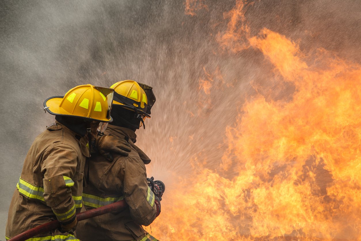 Two firefighters using a hose to battle a blazing fire, highlighting occupational toxin exposures faced by first responders in the line of duty.