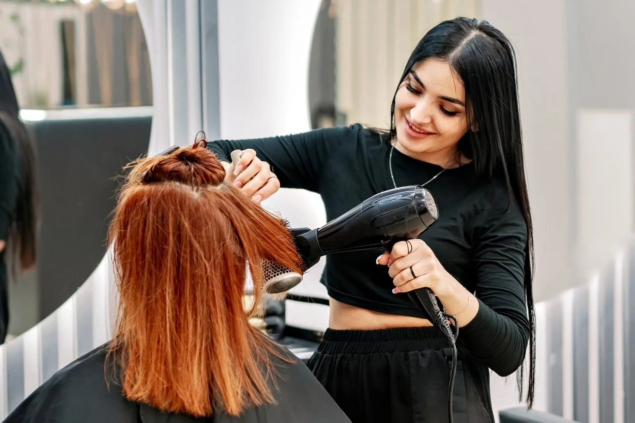 A professional hair stylist blow drying a client's hair in a salon, highlighting workplace chemical and fragrance exposures that can affect bladder health..