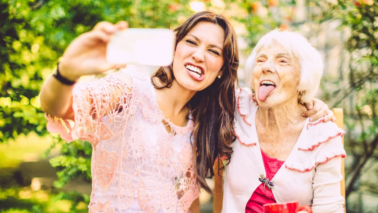 A joyful older woman sticking her tongue out while taking a selfie with a young woman, celebrating generational health equality and vibrant longevity.