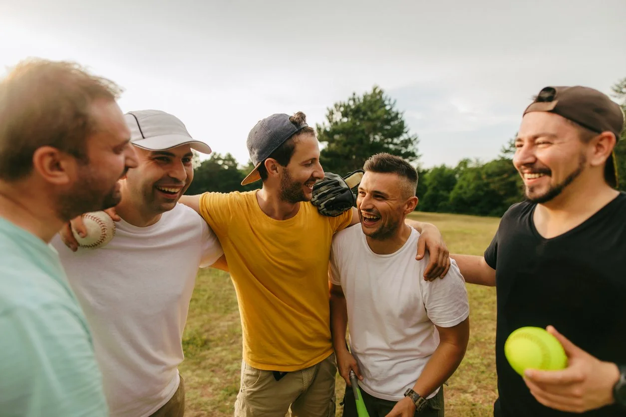 A diverse group of adult male friends laughing together outdoors, normalizing positive men's health, genuine connection, and community across all ages.