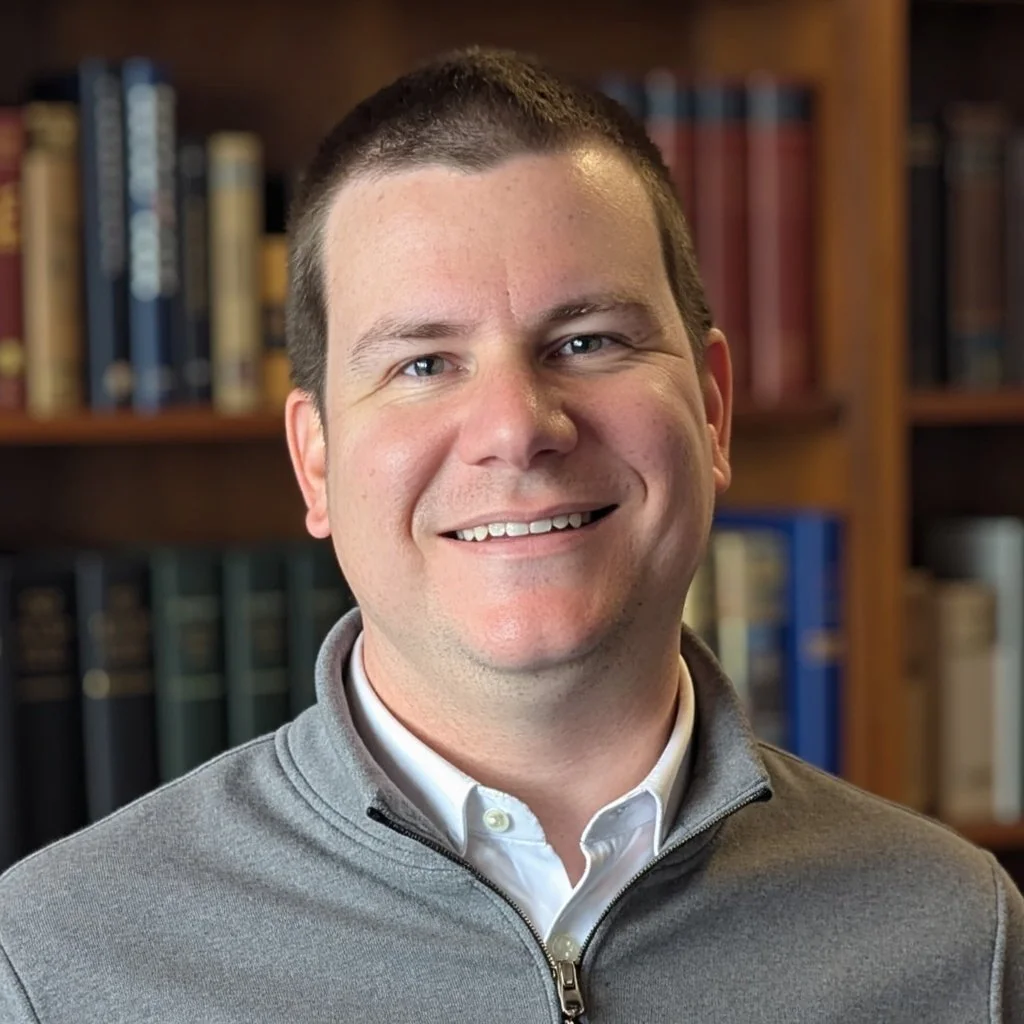 Logan Metesh smiling, wearing a light gray zip-up jacket and a white collared shirt, with bookshelves in the background.