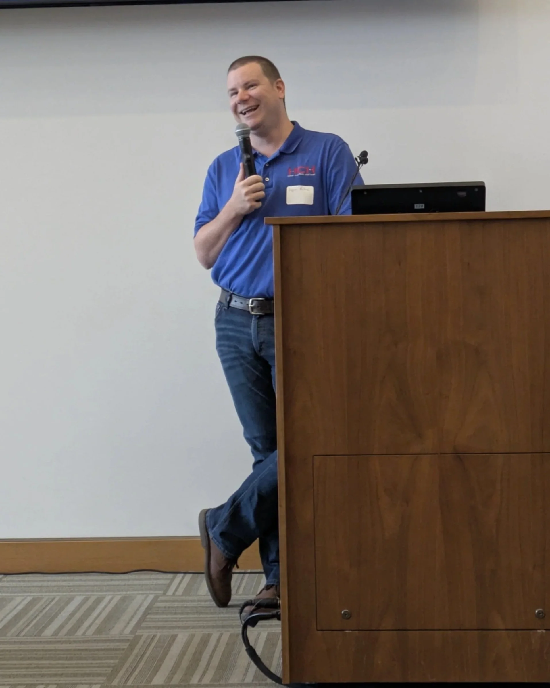 Logan Metesh in a blue polo shirt with a name tag, standing behind a podium with a laptop, holding a microphone, and smiling, in a conference room.