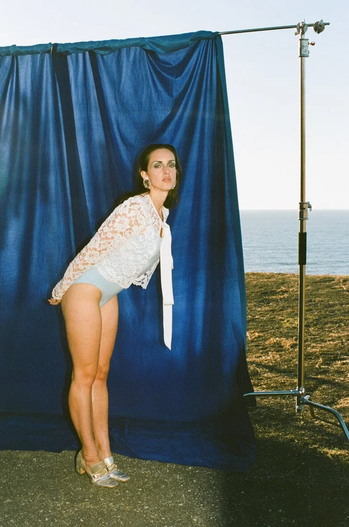 Fashion model with dark hair wearing a lace top, high-waisted shorts, and high heels, posing in front of a blue curtain outdoors with the ocean in the background.
