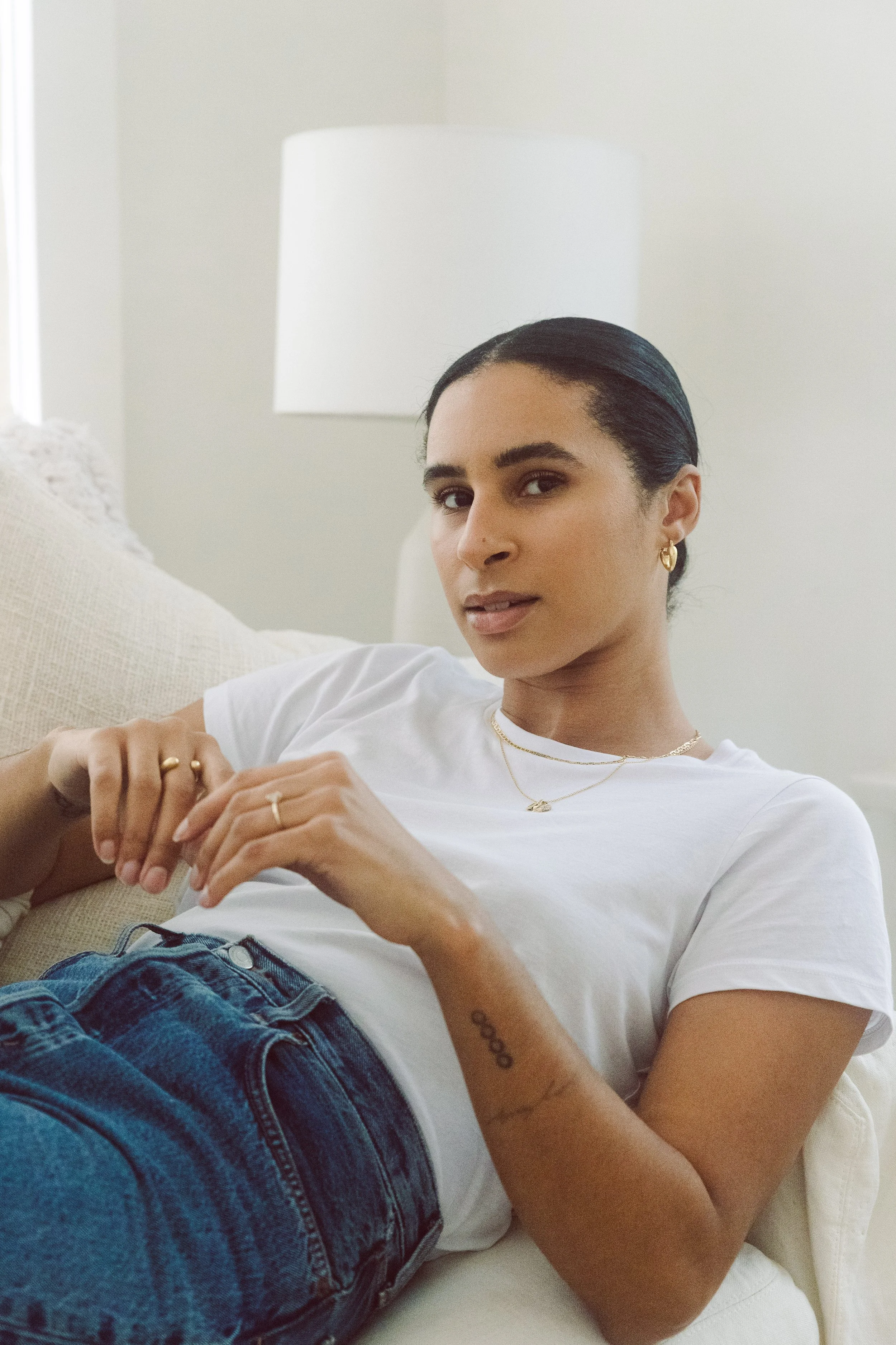 Model in a white T-shirt and jeans seated on a sofa in a bright interior.
