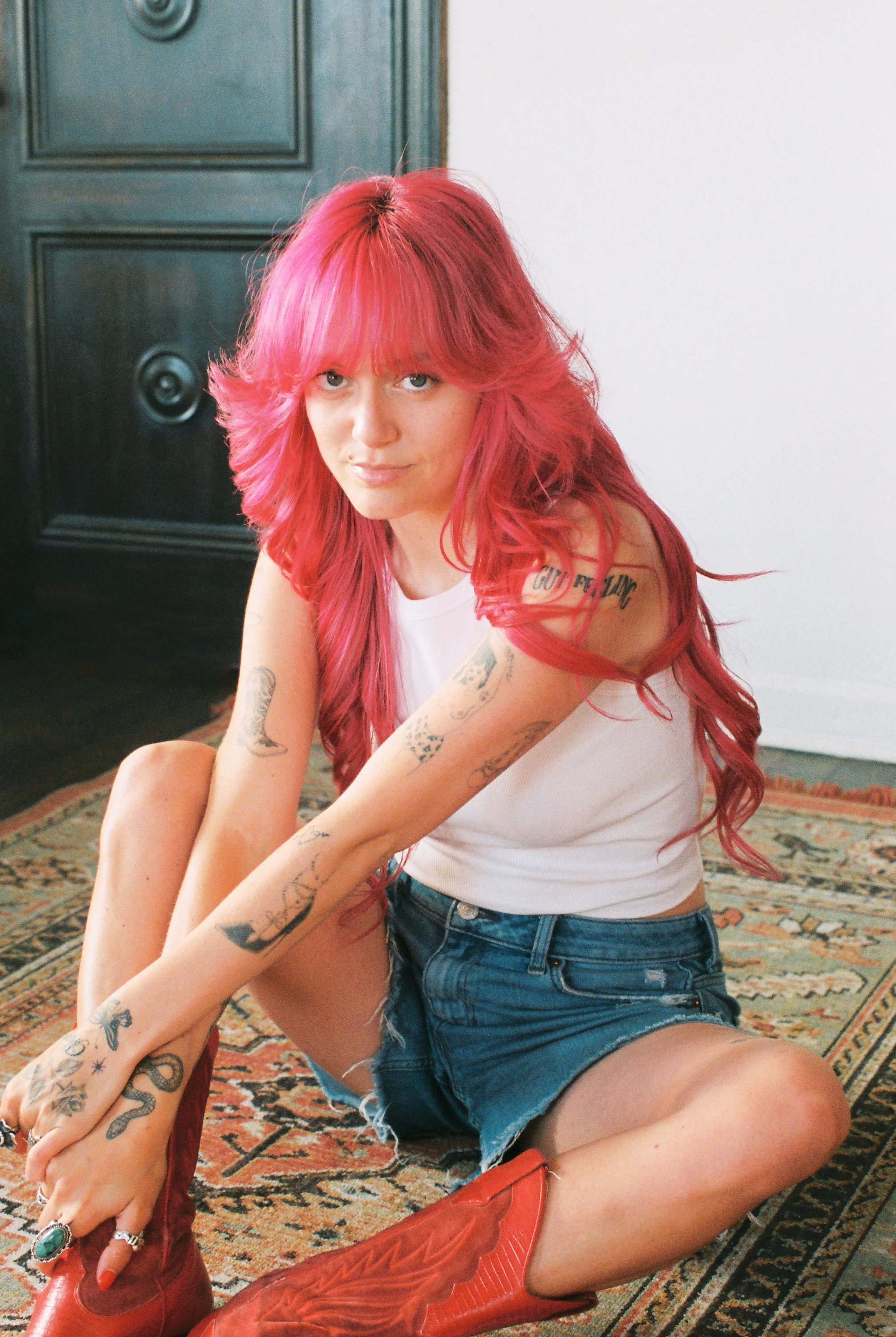 Florence Given, with bright pink hair and tattoos wearing an orange cowboy hat, white tank top, denim shorts, and red cowboy boots, sitting on the floor against a white wall and black door.