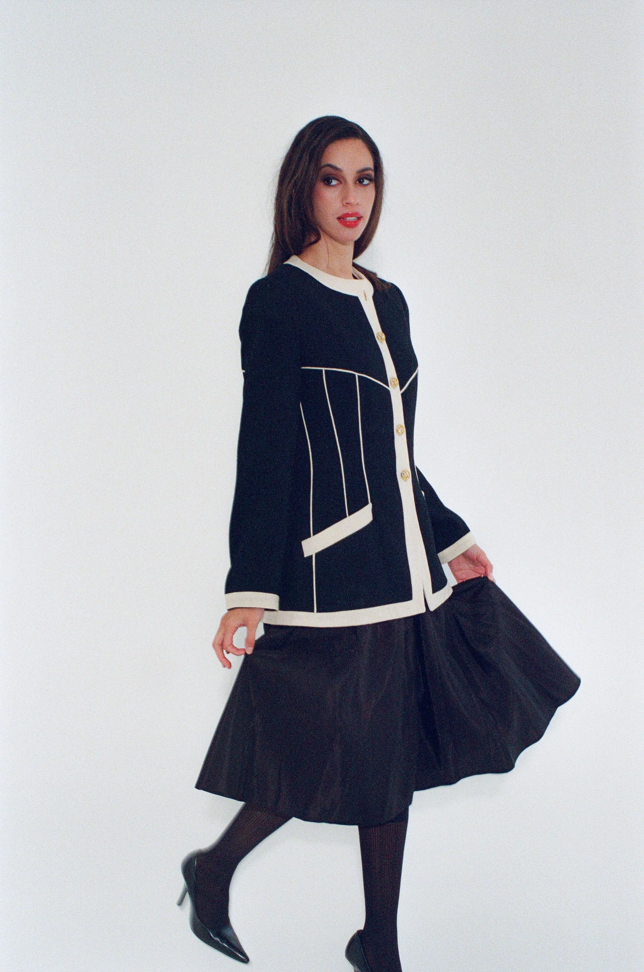 Fashion model with dark brown hair wearing a black and white blazer and a black pleated skirt, standing against a white studio background.