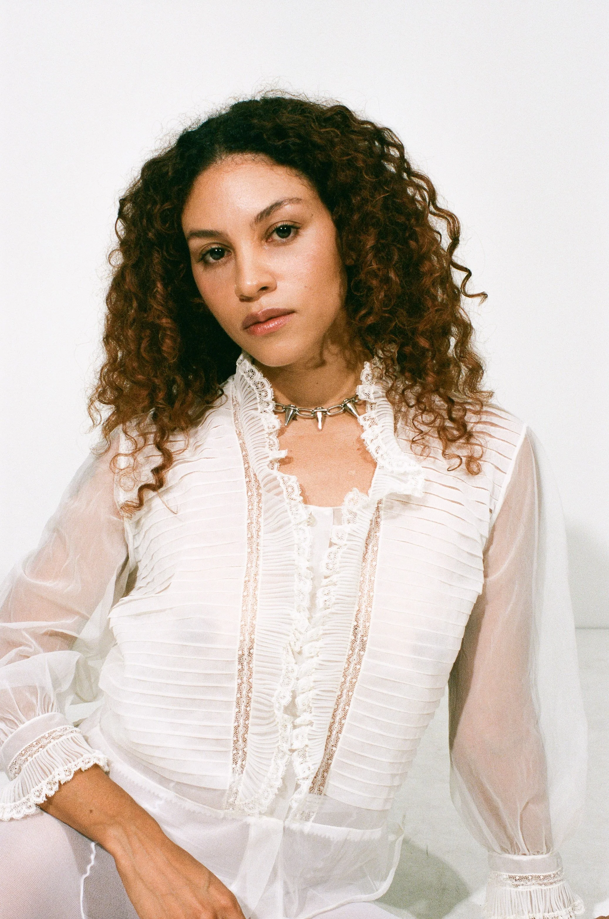 Fashion model with curly hair wearing a white blouse with lace and pleated details, a silver choker necklace, and white pants, against a white studio background.