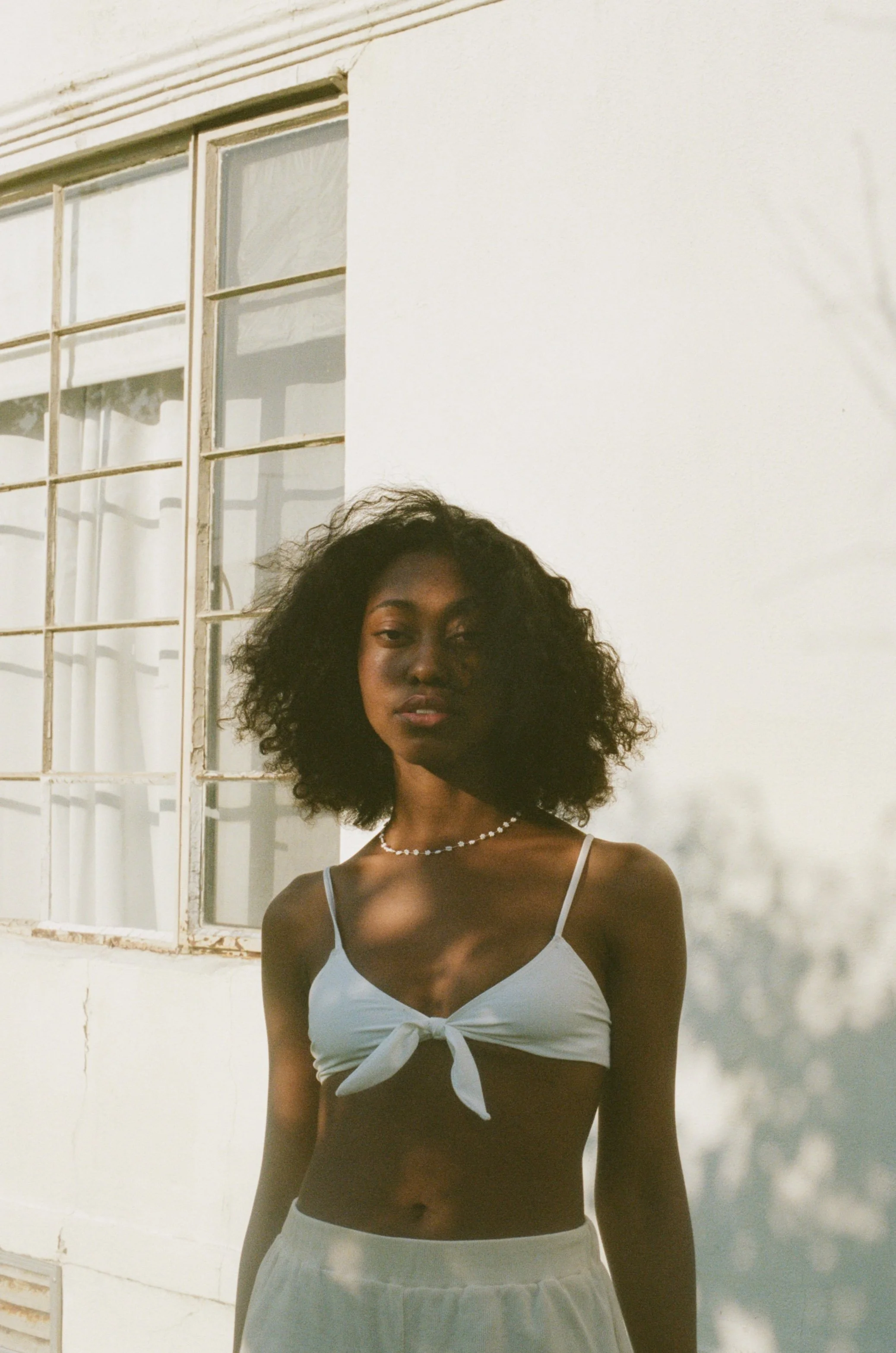 A young woman with curly hair wearing a white crop top with a bow in the front and a string of pearls, standing outside in front of a white wall with a window.