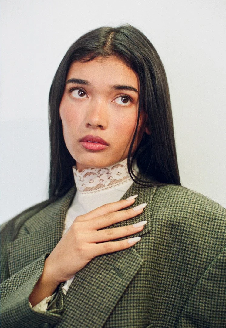 Fashion model with long dark hair wearing a plaid blazer and a white lace turtleneck, posing against a light background.