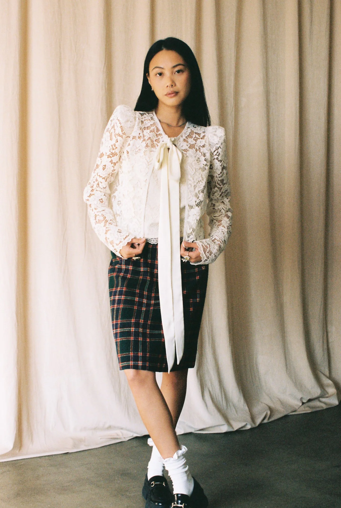 A woman with long black hair standing in front of beige curtains, wearing a white lace blouse with a bow tie, a plaid skirt, white socks, and black platform shoes.