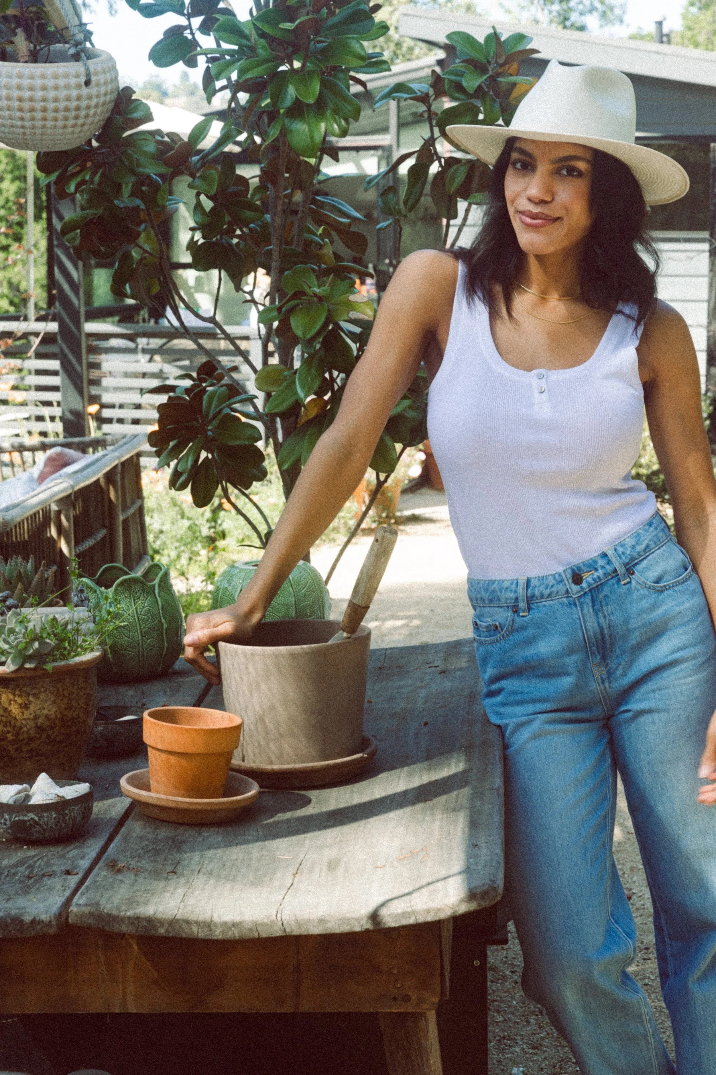 A model wearing a white hat and sleeveless white top standing outdoors next to a wooden table with potted plants, including succulents and leafy green plants, in a garden or backyard setting.