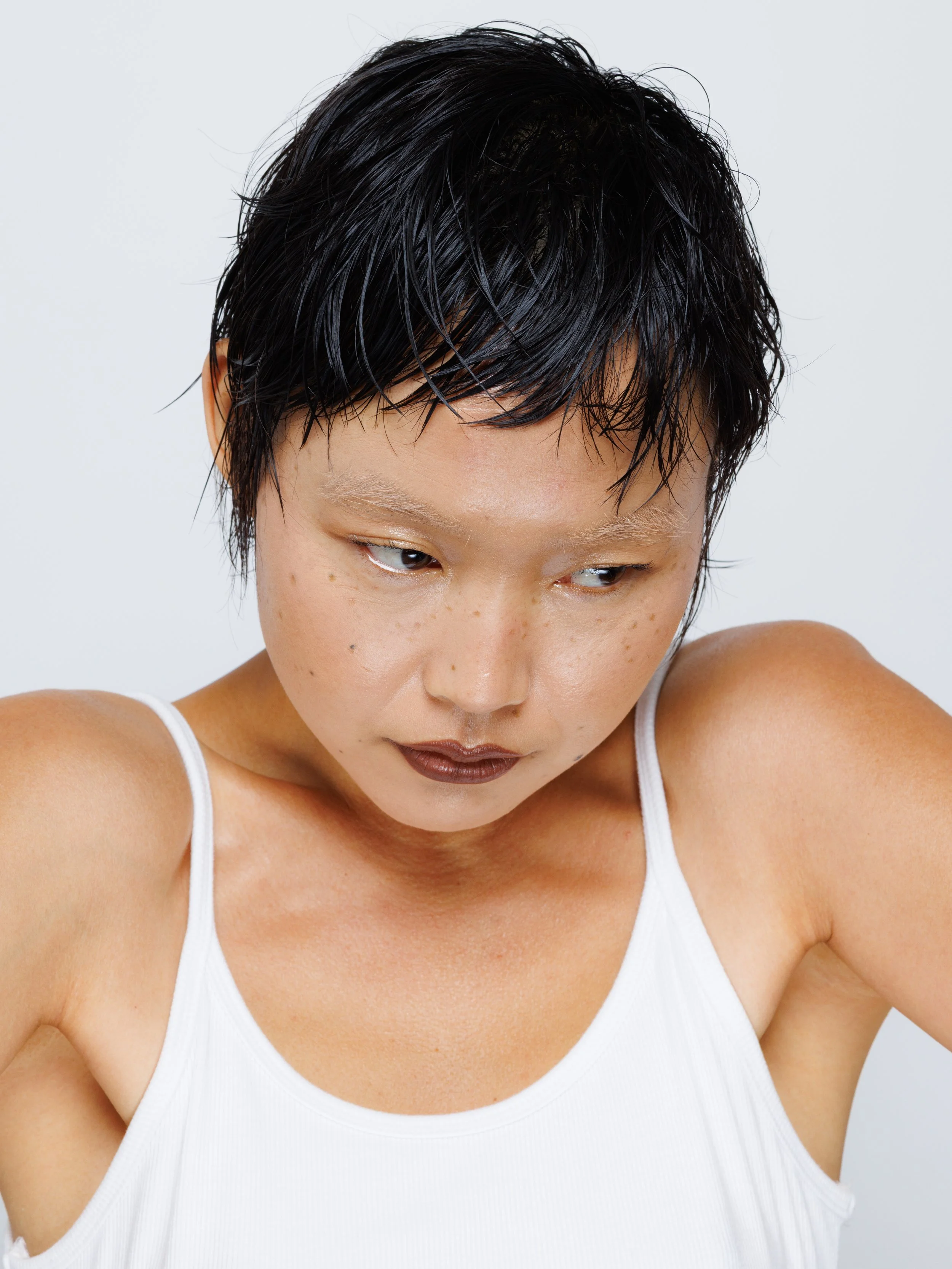 A person with short black hair looking downward, wearing a white tank top, with a neutral background.