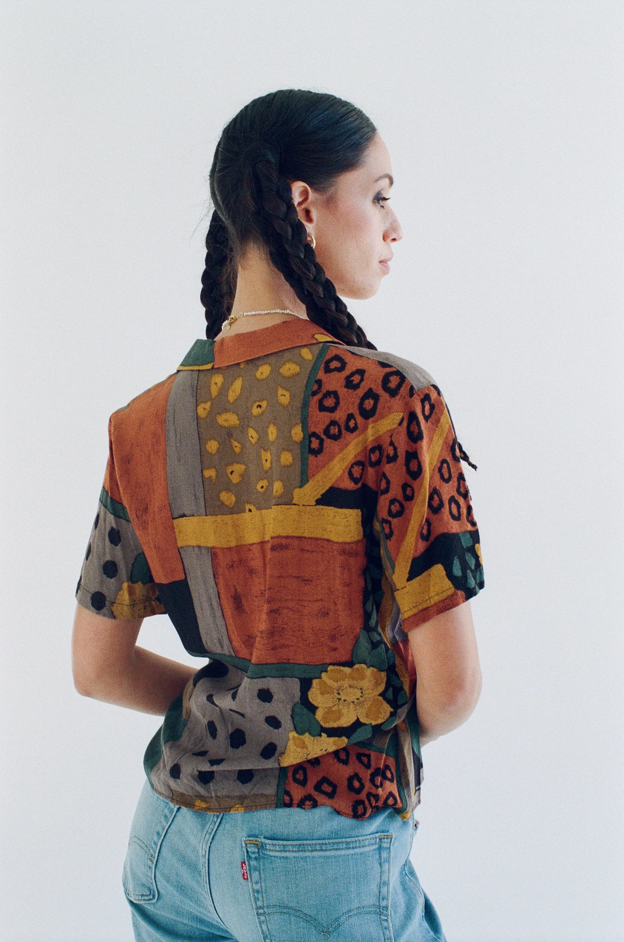 Fashion model with black hair in two braids wearing a colorful patterned short-sleeve shirt and light blue jeans, standing against a white background and facing right.