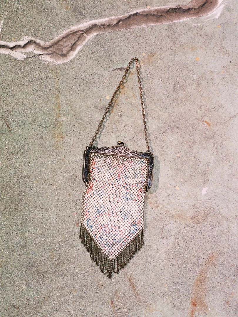 Antique evening bag with a chain strap, rhinestones, and fringe hanging against a textured concrete wall.