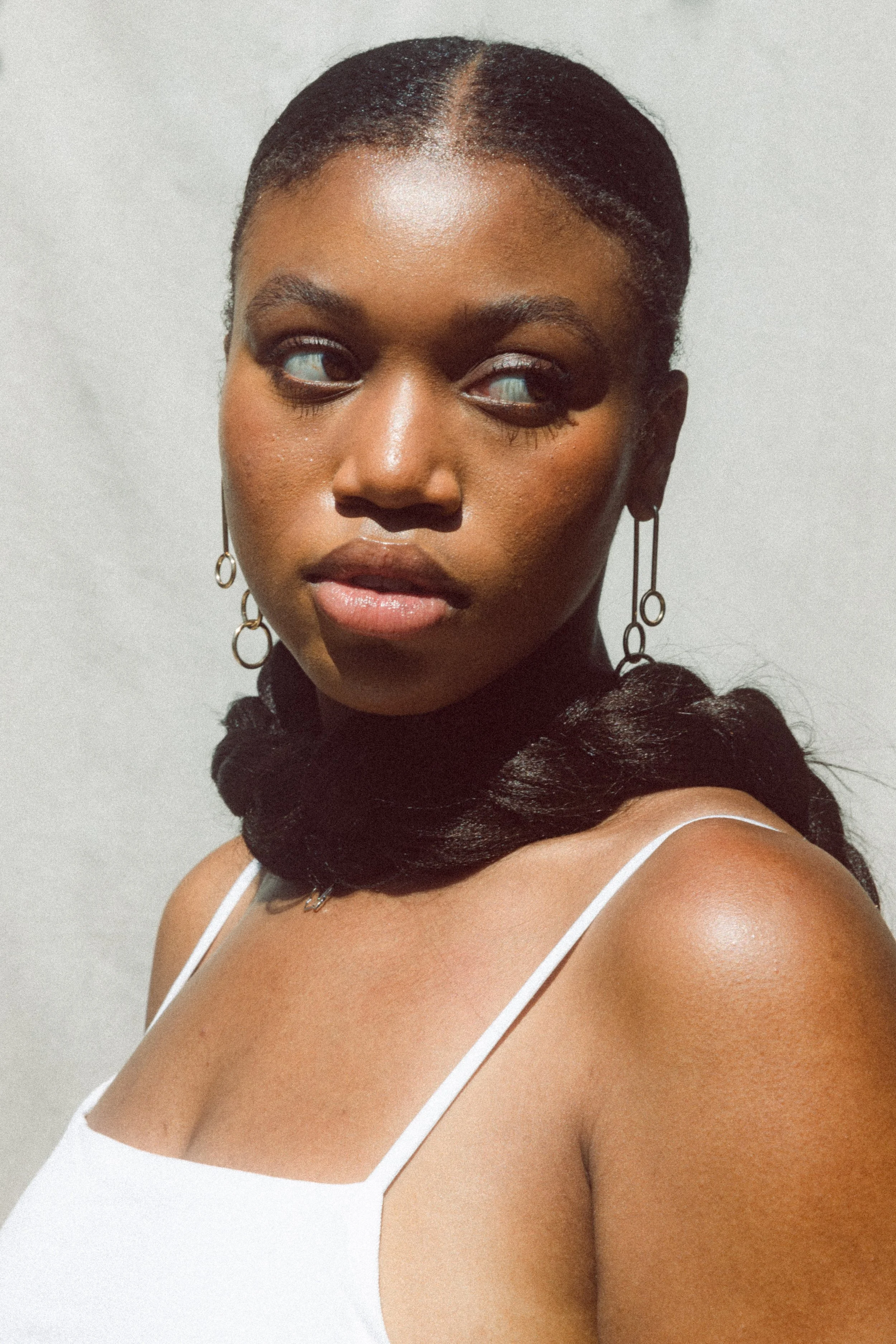 Close-up of a woman with dark skin and braided hair, wearing earrings, a black scarf around her neck, and a white tank top, with a neutral background.