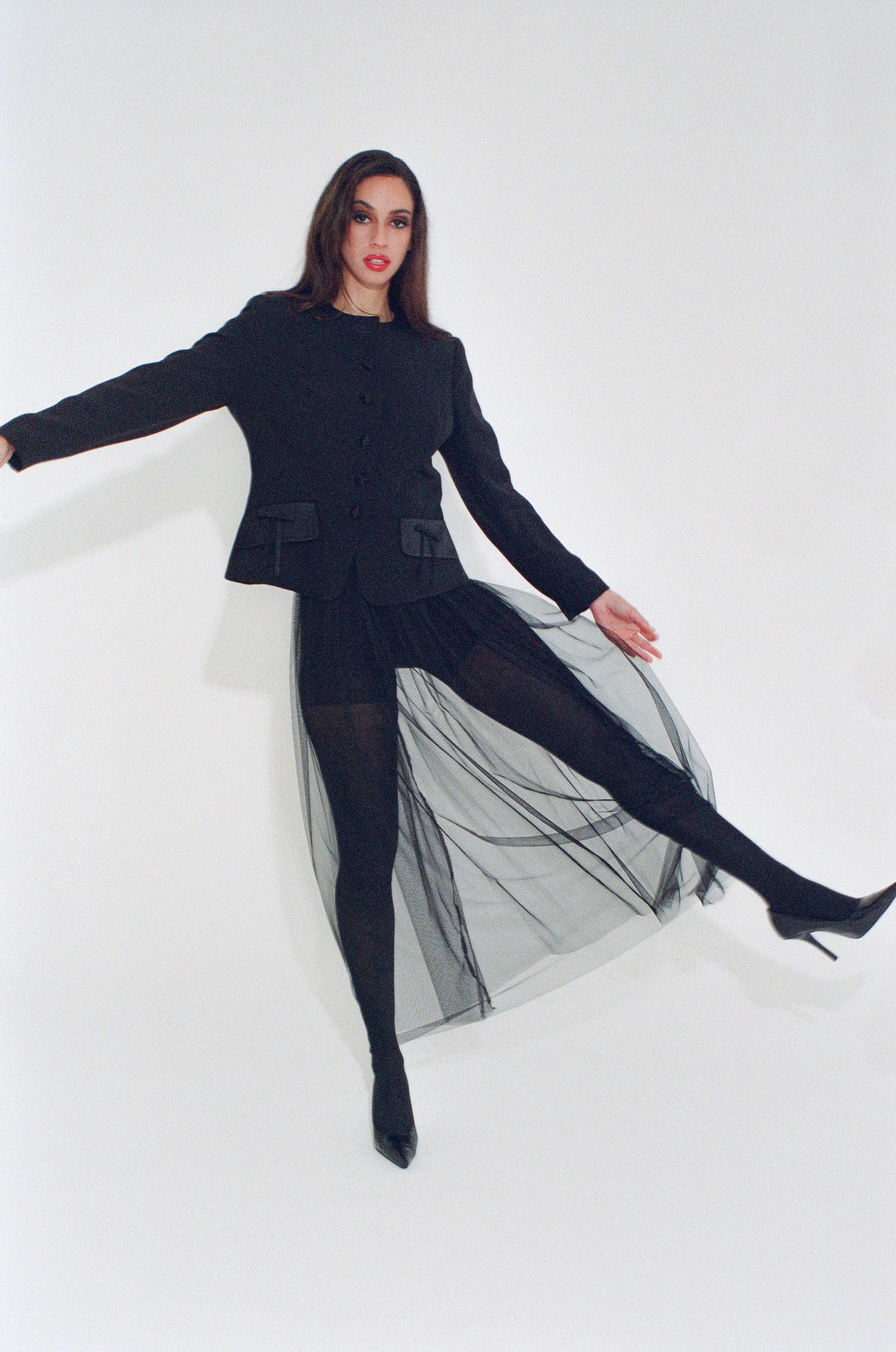 Fashion model with long dark hair wearing a black blazer and a sheer black tulle skirt, posing with her arms outstretched against a light background.