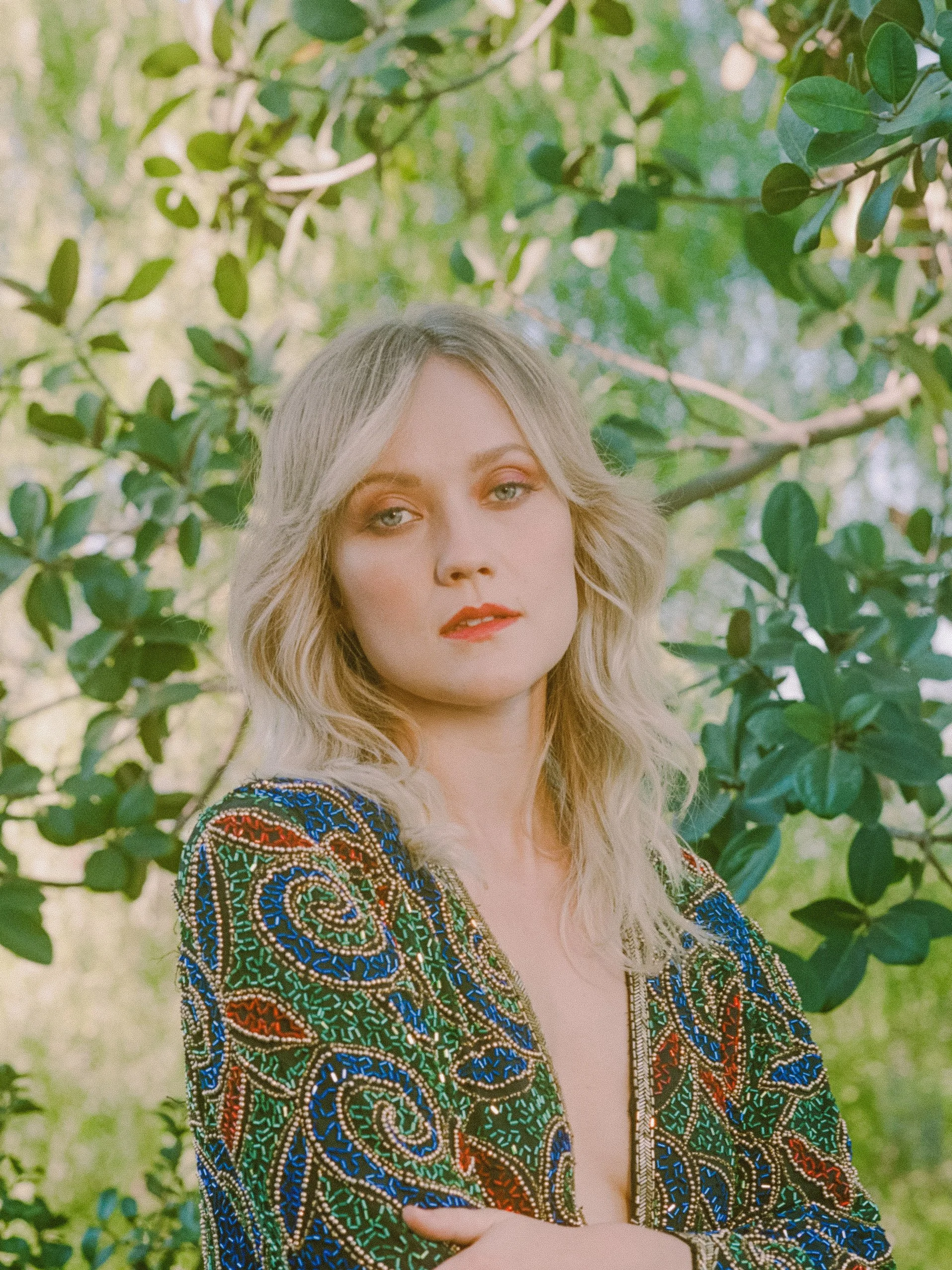 A woman with blonde hair and makeup posing outdoors surrounded by green leaves, wearing a colorful, intricate embroidered jacket.