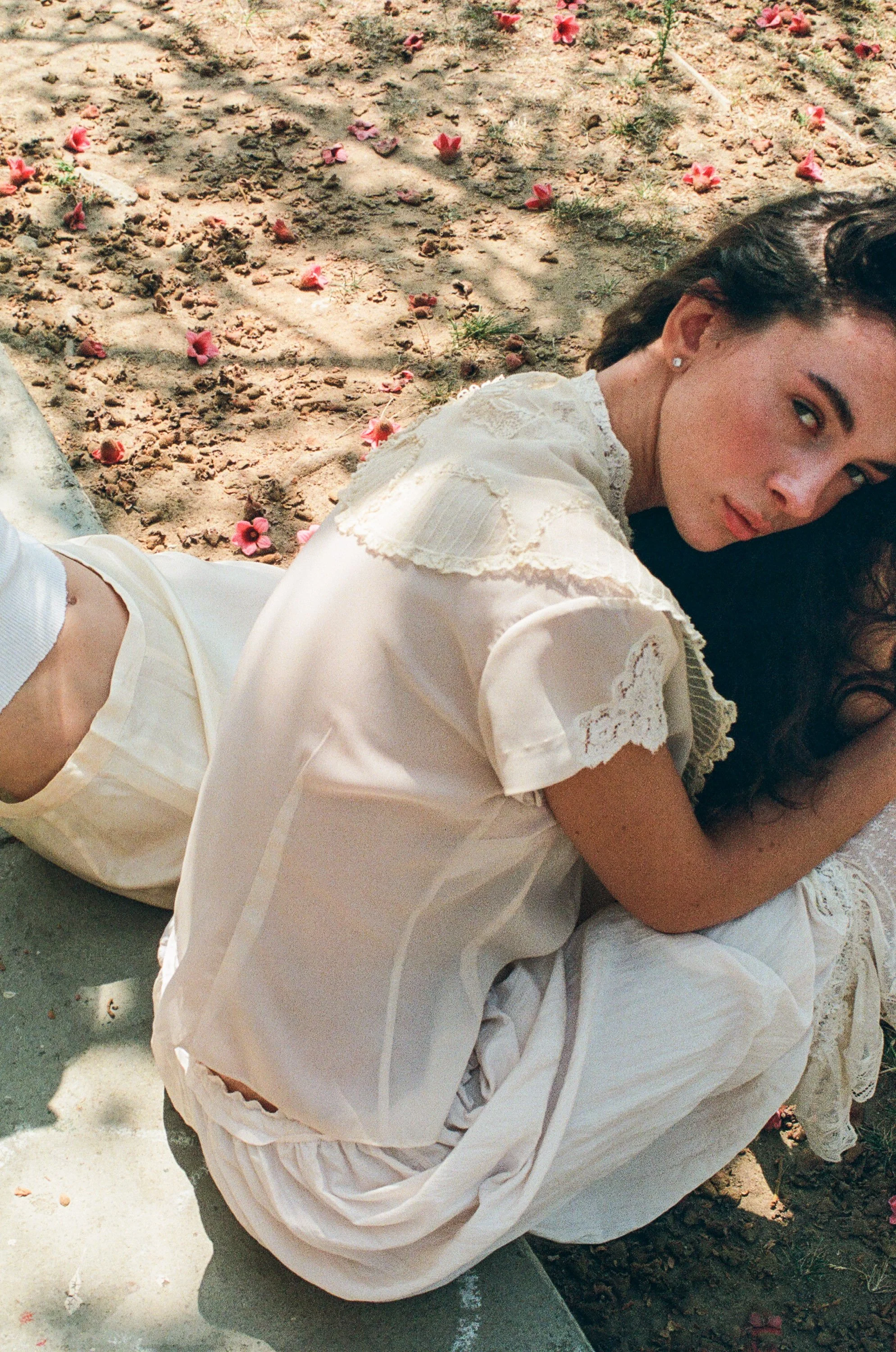 Model with long dark hair lying on her side among pink flower petals, wearing a white lace dress and looking at the camera.