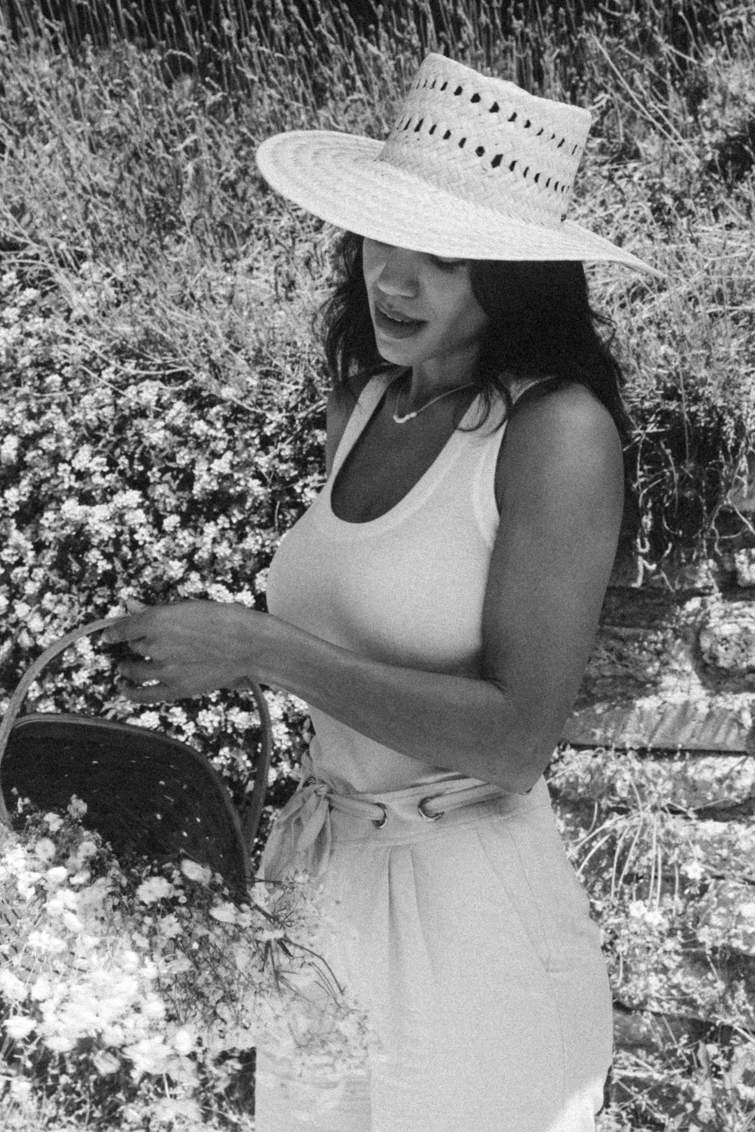 Model in a sun hat and white dress standing among wildflowers in a black-and-white portrait.