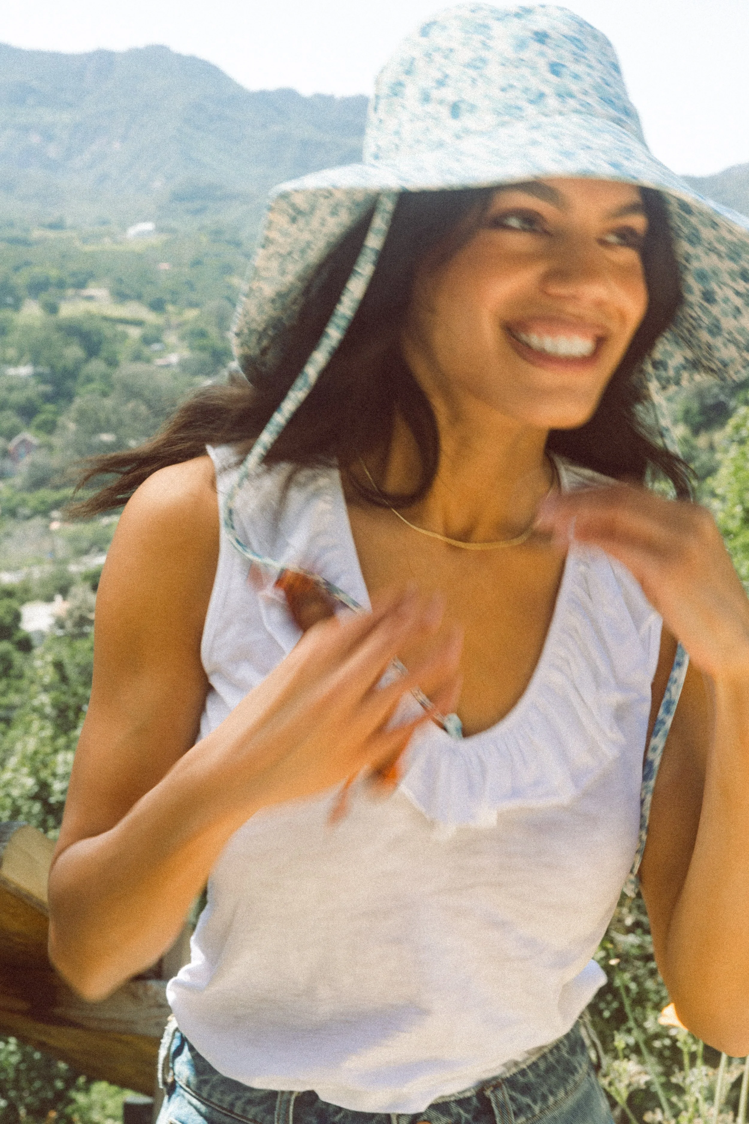 Smiling model in a white tank top and floral sun hat outdoors in a mountainous landscape.