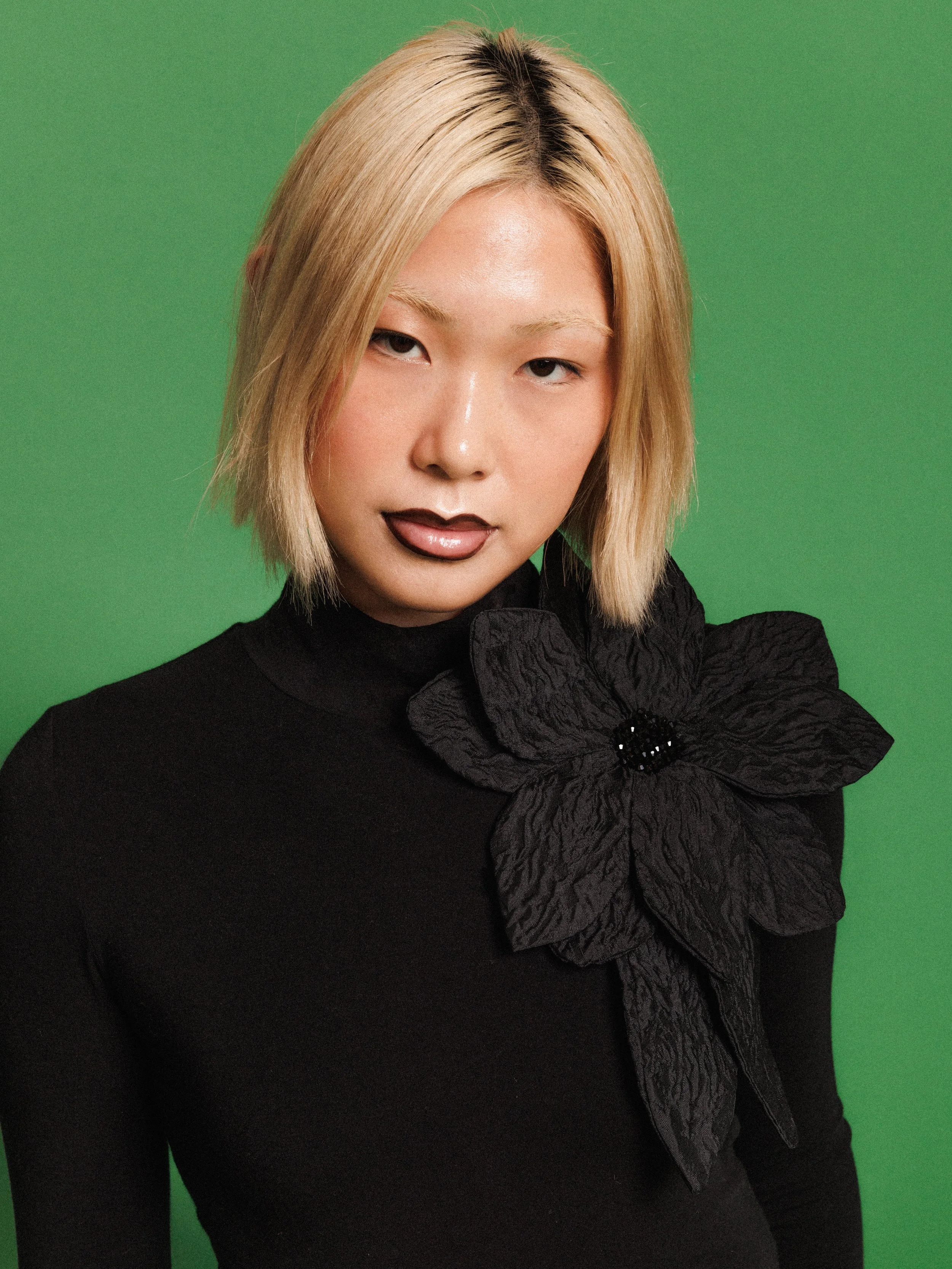 Editorial fashion portrait of a model wearing a black long-sleeve top with an oversized flower, photographed against a vibrant green studio background.