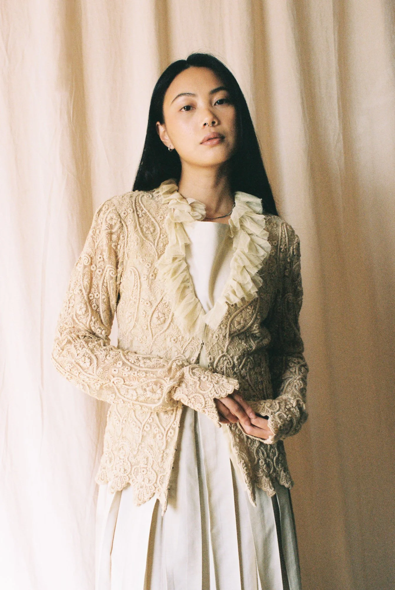 Portrait of a woman with long black hair wearing a cream-colored lace jacket with ruffled collar, standing in front of beige curtains.