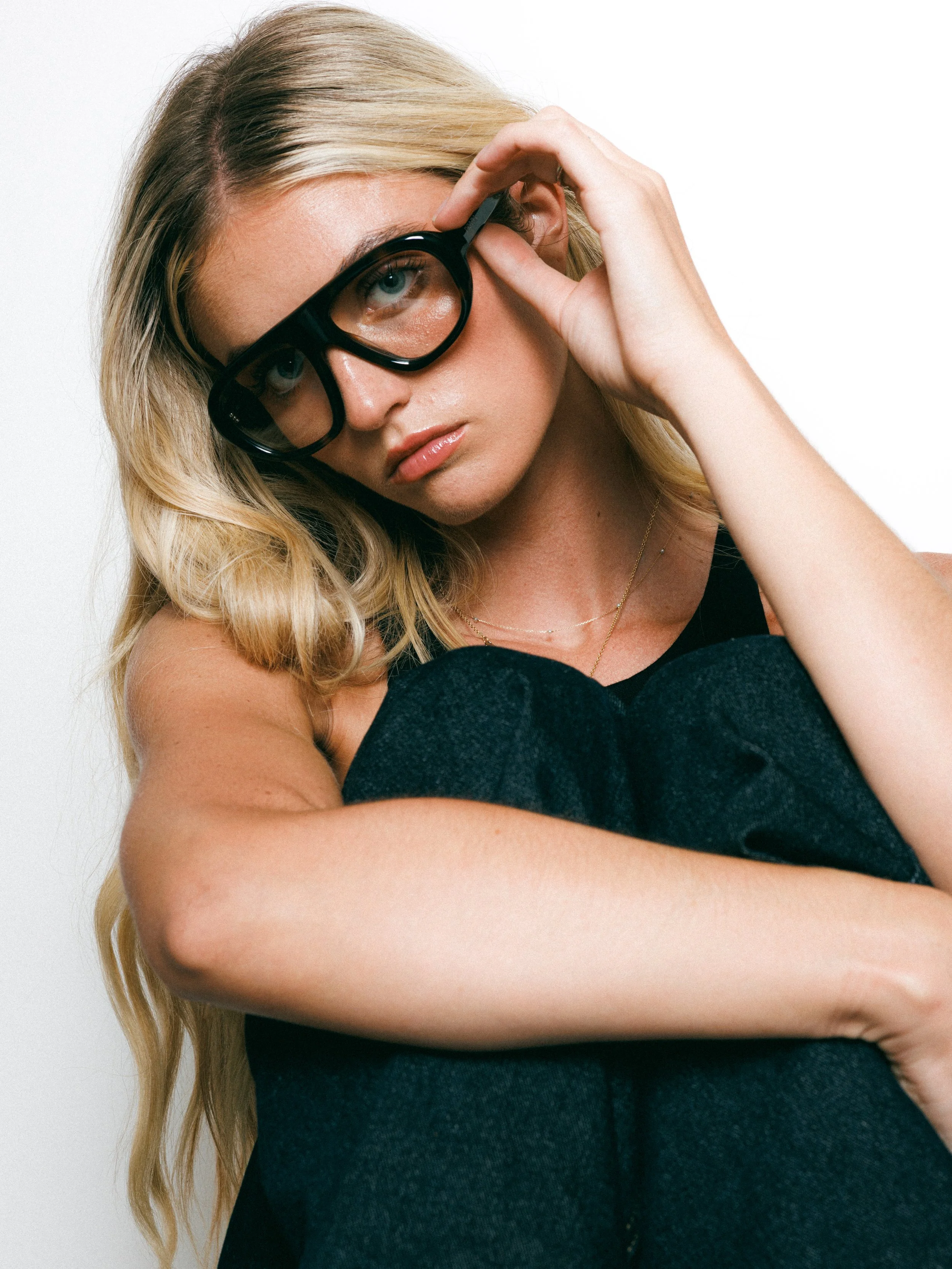 Close-up portrait of a blonde fashion model wearing oversized black glasses, gold hoop earrings, and a black sleeveless top against a white background.