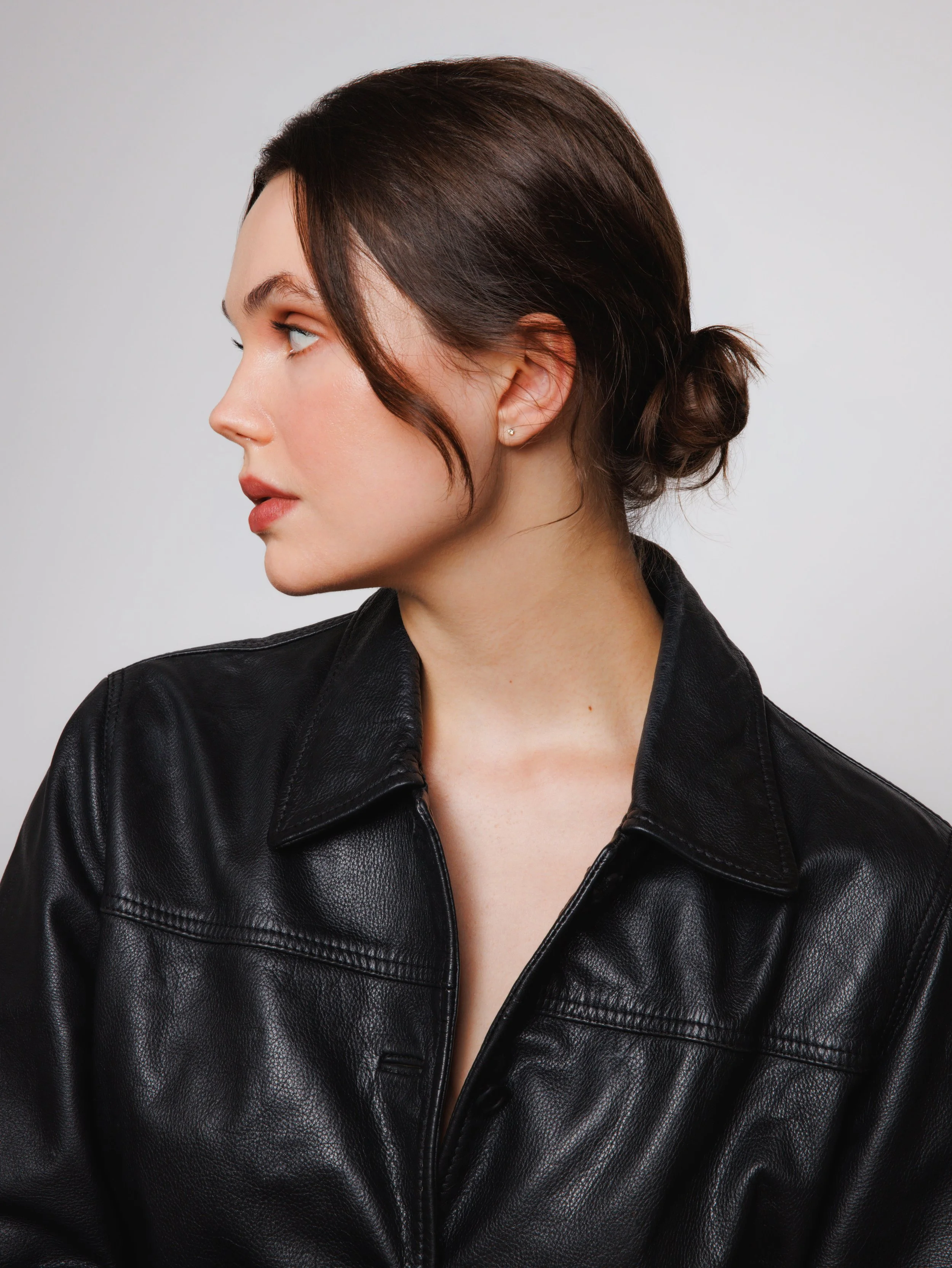Beauty portrait of a model with dark hair styled in a low bun, wearing a black leather jacket and photographed in profile against a clean light gray background.