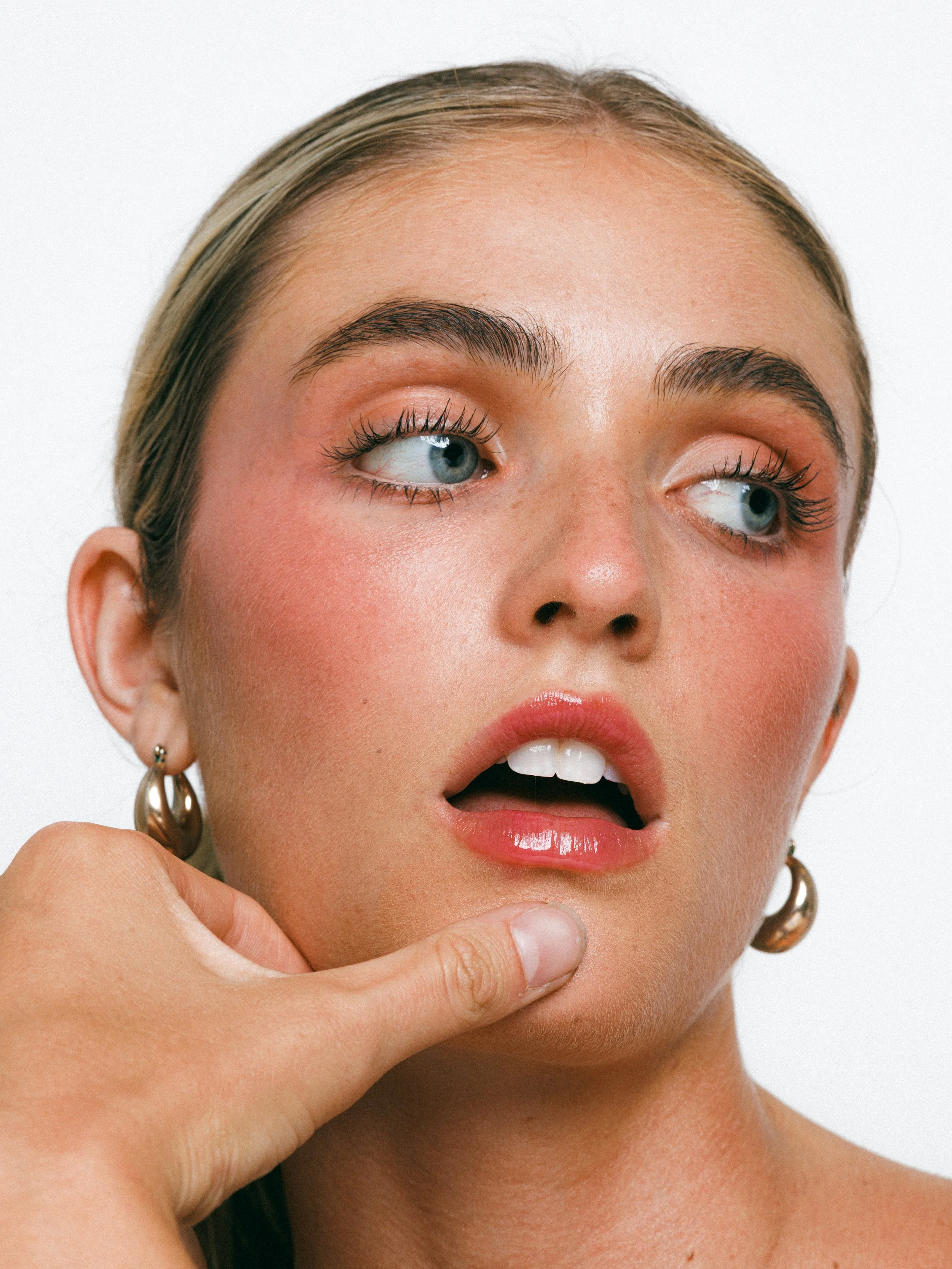 Close-up beauty portrait of a blonde model with glowing makeup, gold hoop earrings, and a sleek ponytail against a white background.