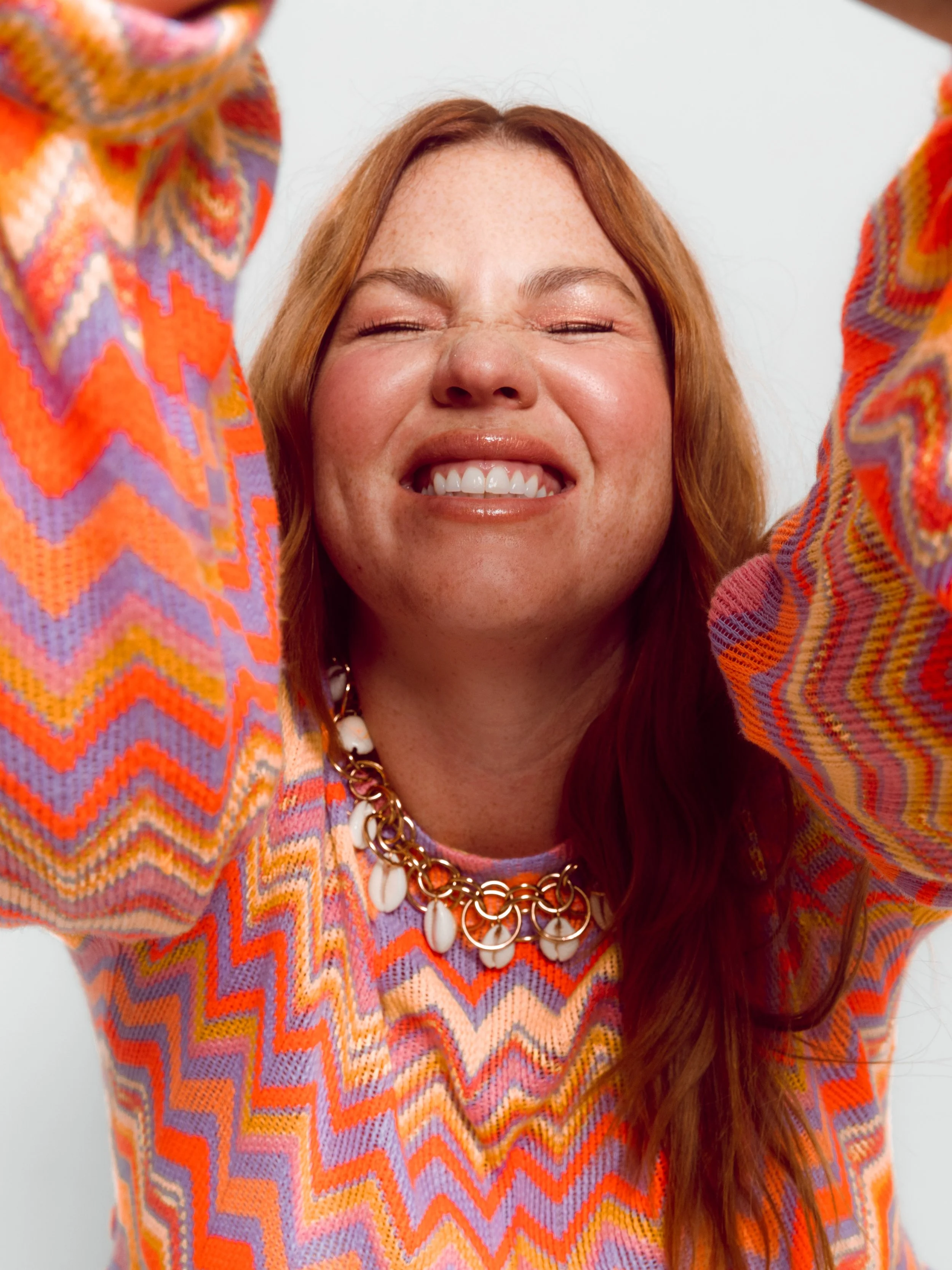 Fashion model with red hair smiling with her eyes closed, wearing a colorful chevron-patterned sweater and a chunky necklace with her arms raised overhead.