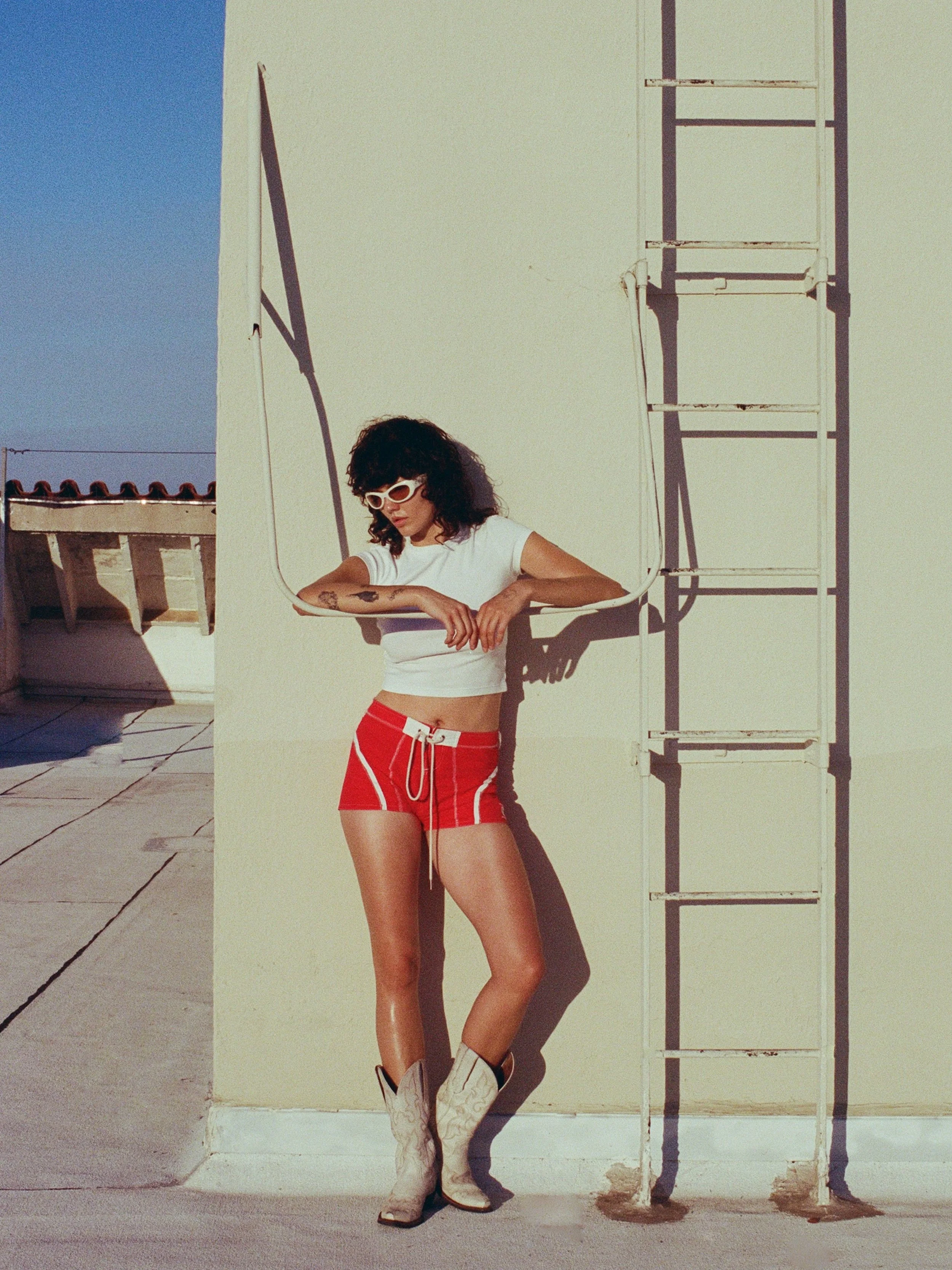 Editorial fashion portrait of a model wearing red shorts, a white top, sunglasses, and white cowboy boots, posed next to an outdoor ladder in bright sunlight.