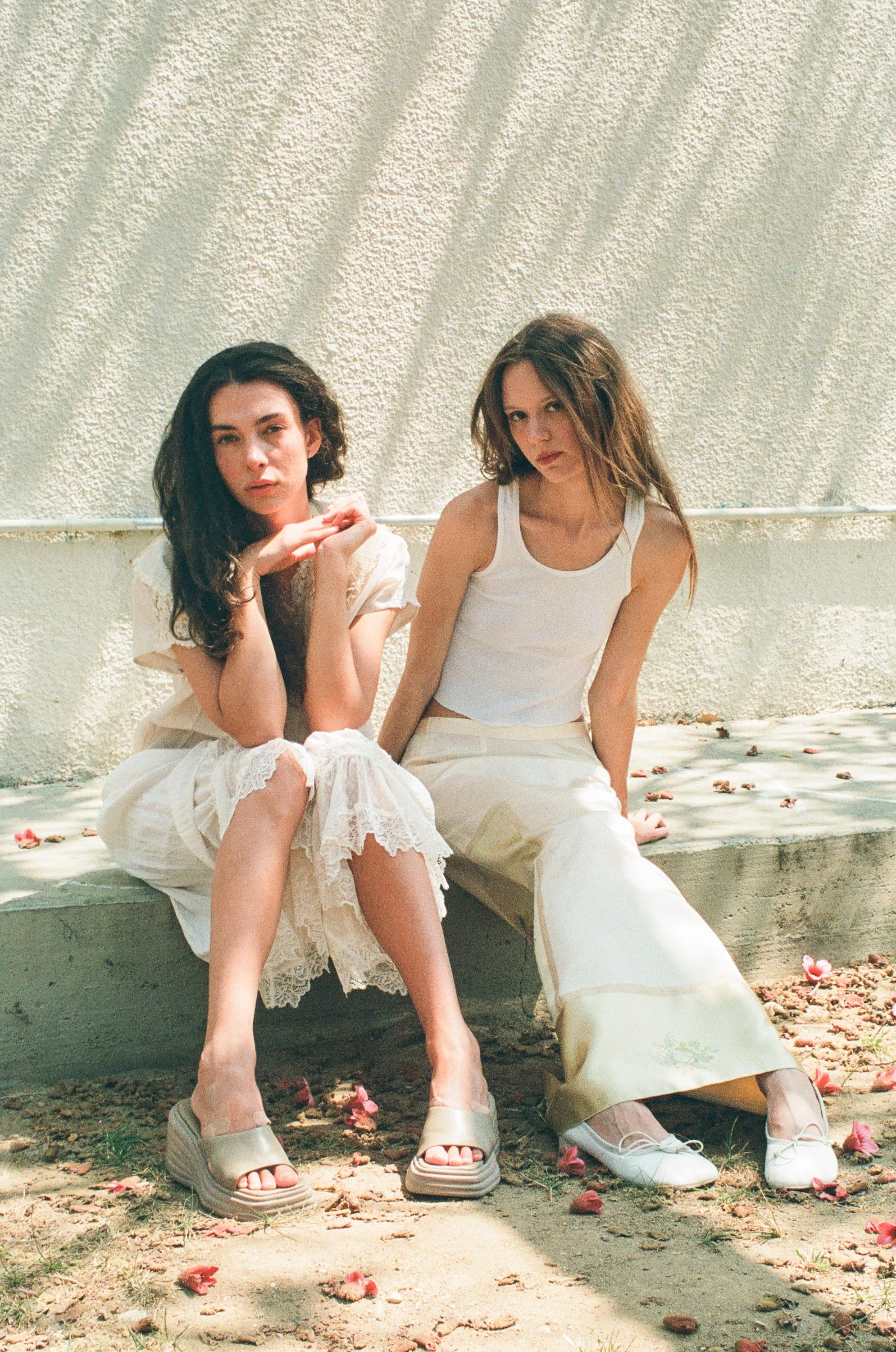 Two fashion models sitting on a concrete ledge against a textured white wall, wearing white and beige clothing with pink petals scattered on the ground.