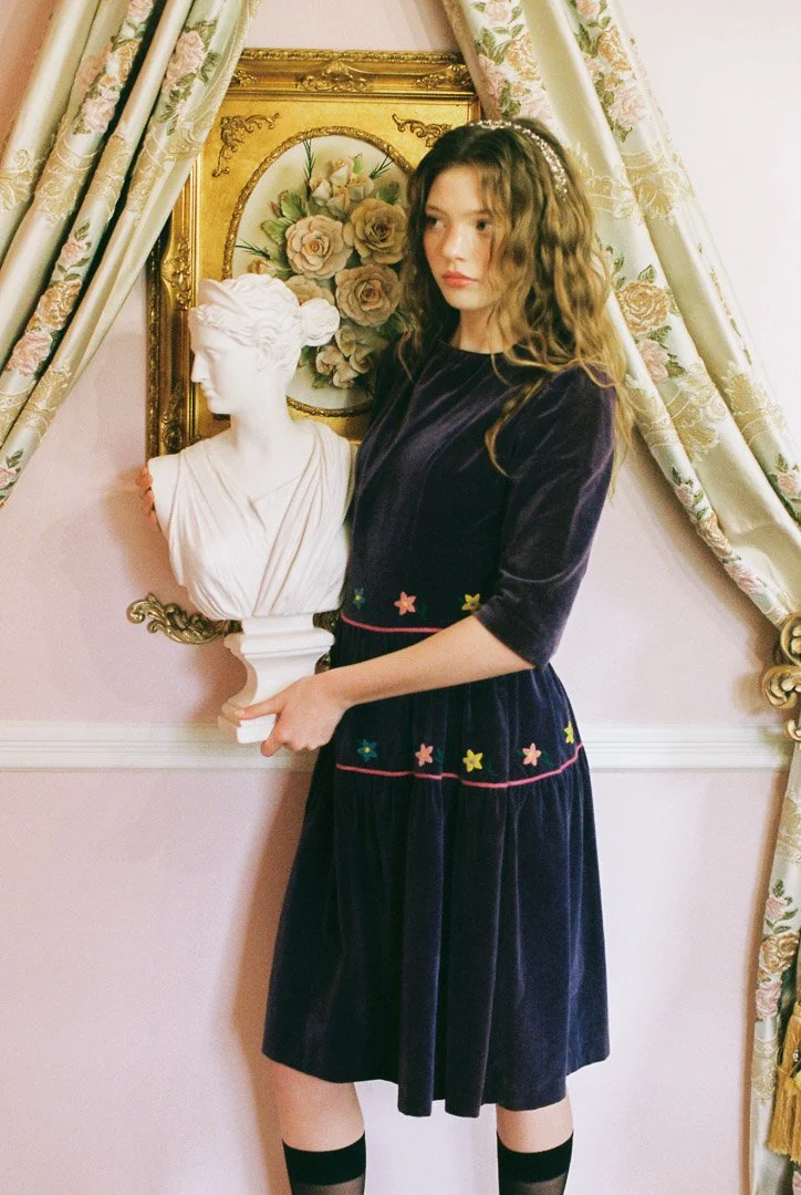 Fashion model with wavy brown hair wearing a dark velvet dress with colorful star embroidery, standing beside a classical white bust in a room with floral curtains and framed floral artwork.