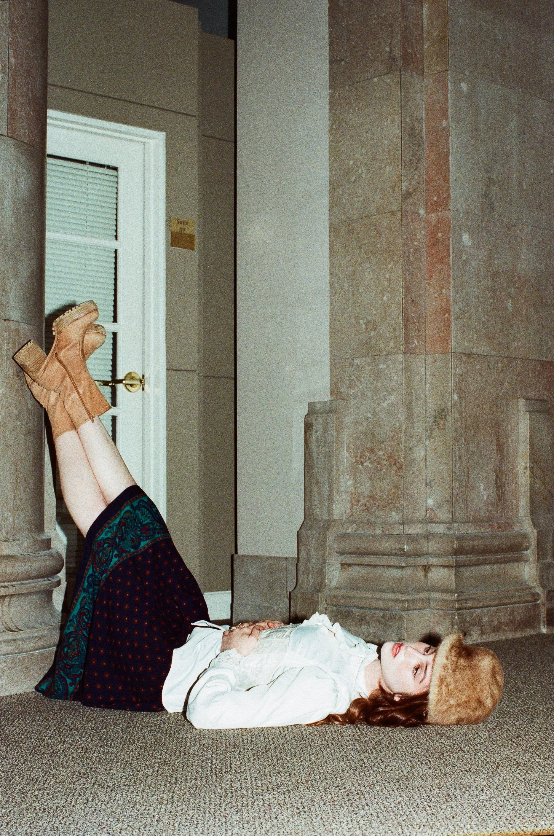 Fashion model with long brown hair lying on the floor of a stone building with her eyes closed, wearing a white outfit as a lion gently paws her cheek.