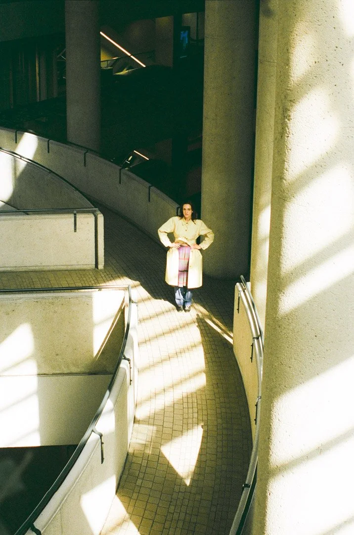 Fashion model wearing a beige trench coat, pink plaid skirt, and dark shoes, standing on a curved tiled walkway in a modern building with sunlight casting dramatic shadows.