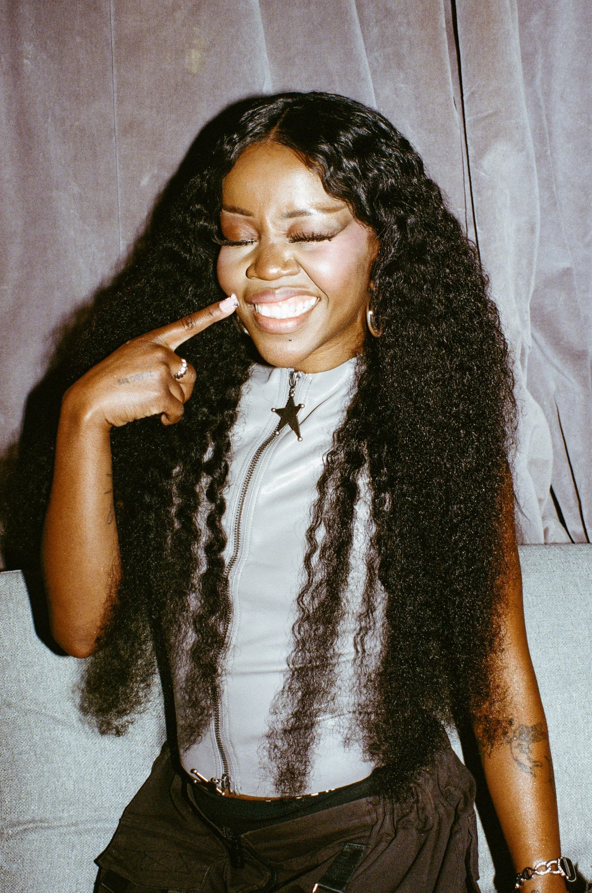 A woman with long, curly black hair smiling with eyes closed, pointing to her cheek with her finger, wearing a white zip-up top with a star necklace, black pants, and jewelry.