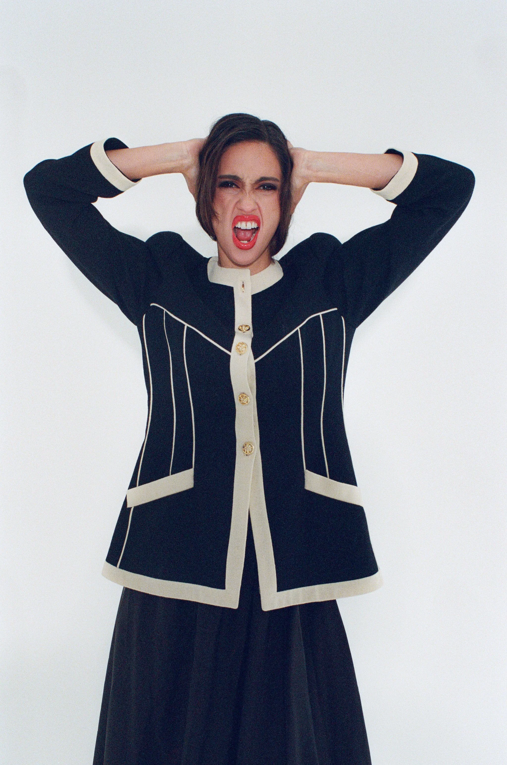 Fashion model with short dark hair wearing a black and white blazer with gold buttons, standing against a white background with her hands behind her head and an expressive pose.