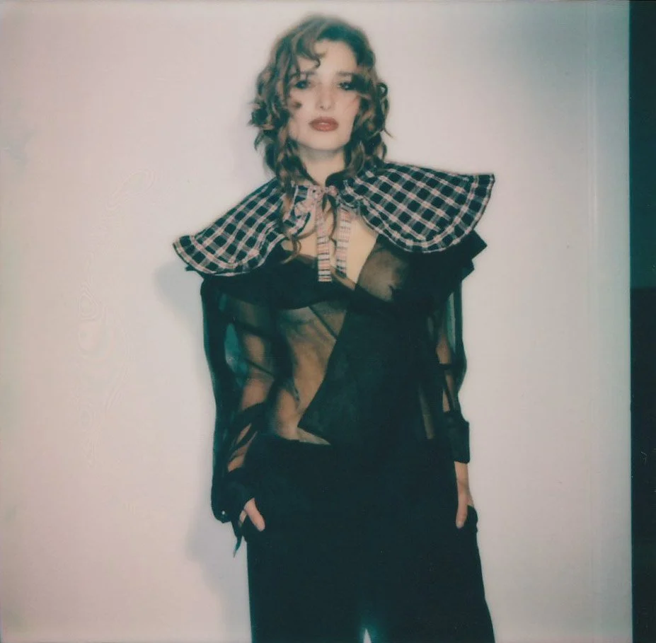 Fashion model with curly hair wearing a sheer black top and a checkered shawl, standing against a plain background.
