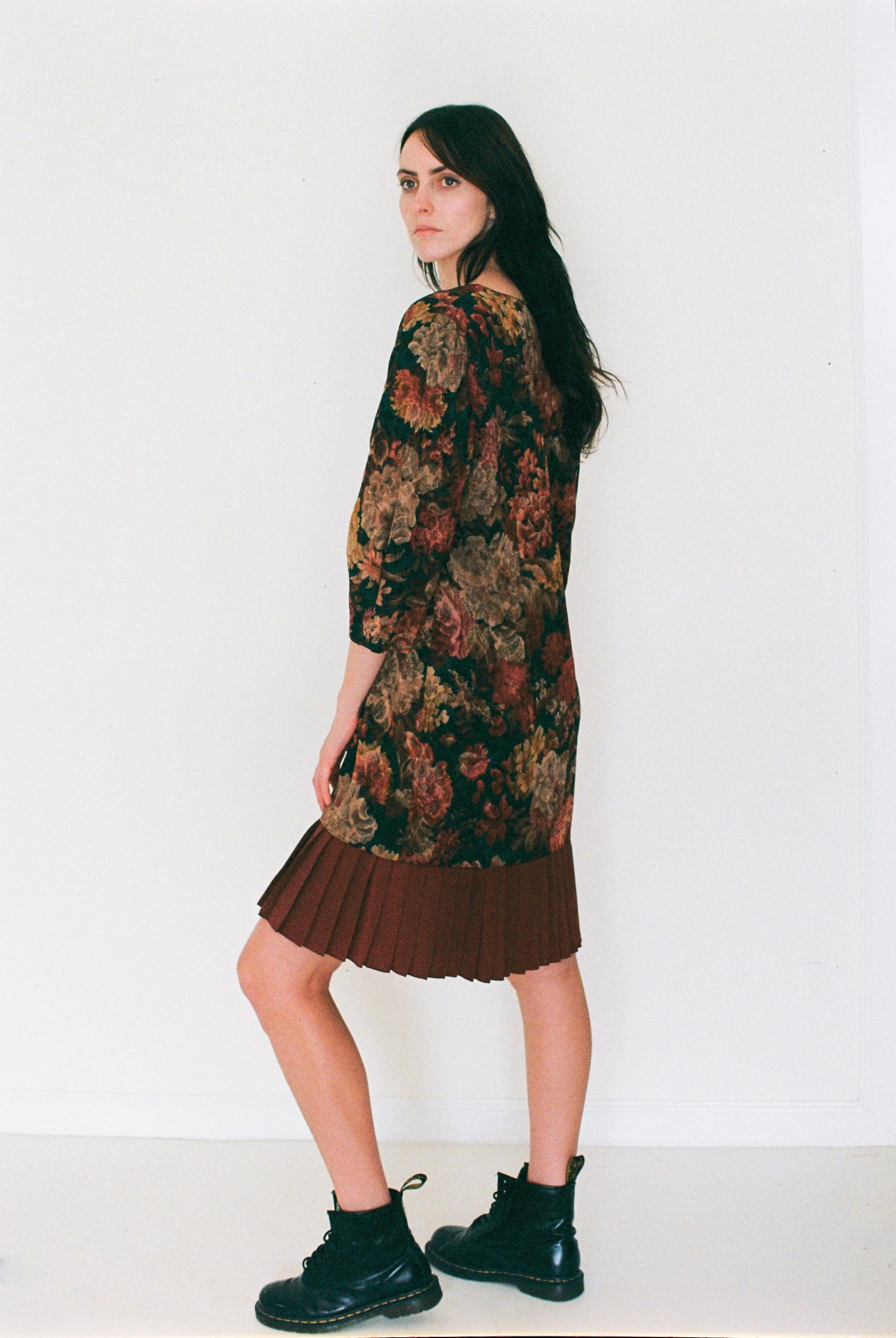 Fashion model standing against a white wall wearing a floral long-sleeve dress, a pleated brown skirt, and black combat boots.