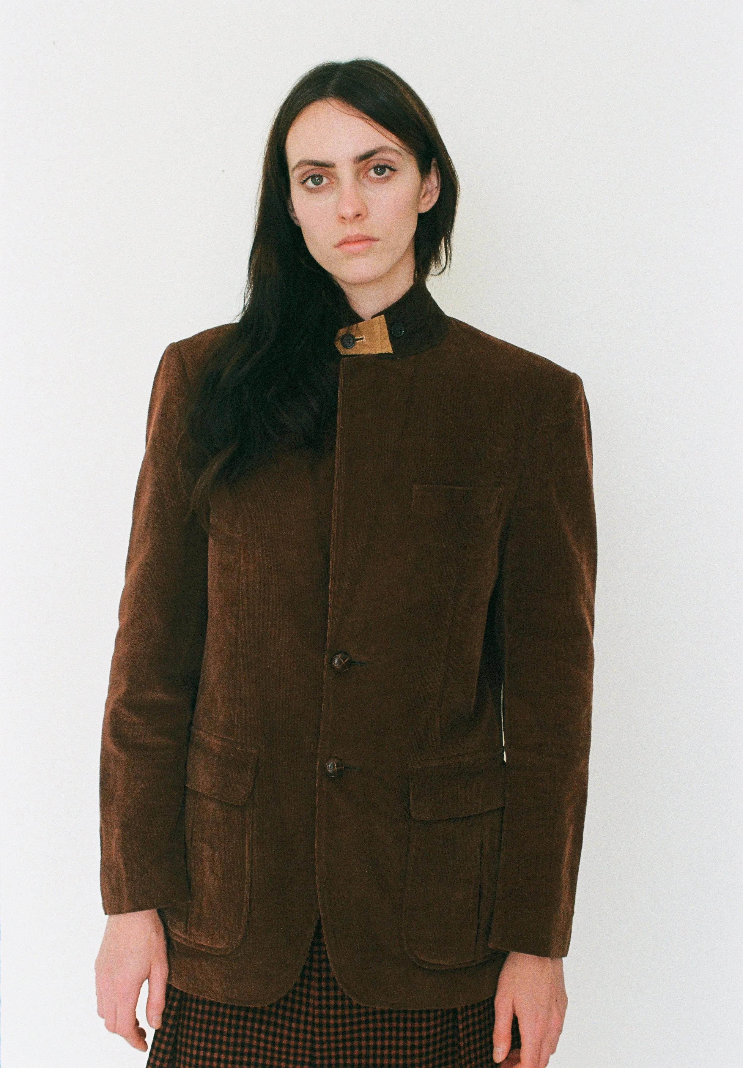 Fashion model with long dark hair wearing a brown jacket and a checkered skirt, standing against a white background.