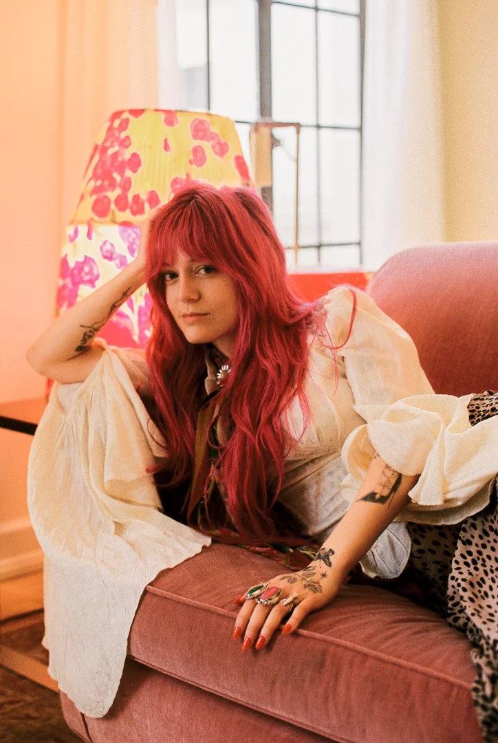 Editorial lifestyle portrait of Florence Given with vivid red hair reclining on a pink sofa, wearing a flowing white outfit in a colorful, warmly lit interior.