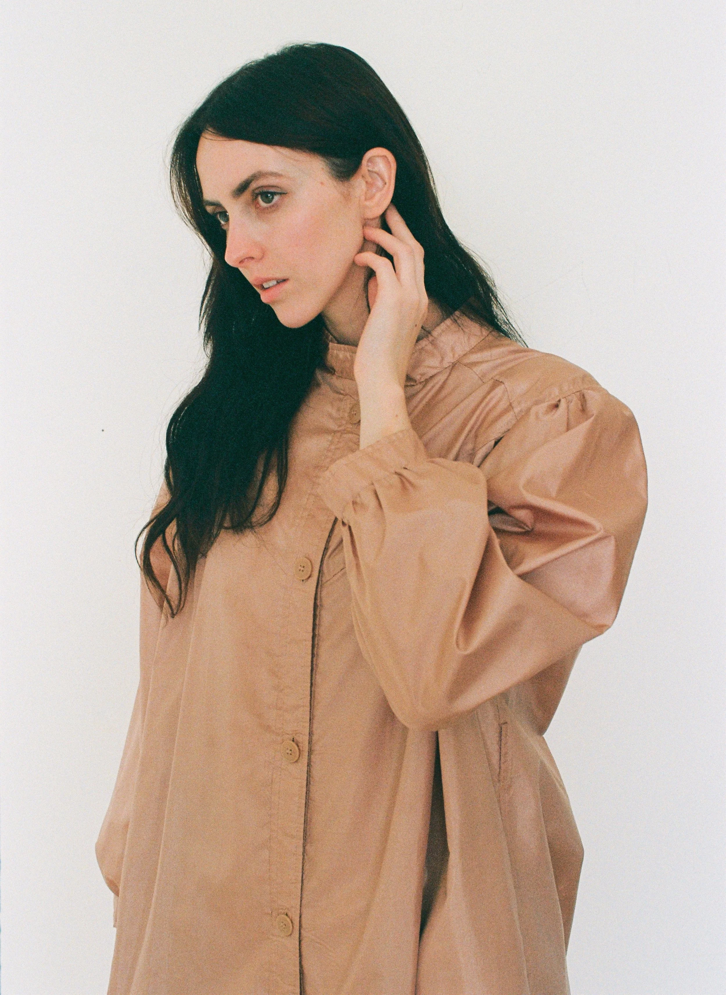 Fashion model with long dark hair wearing a tan button-up jacket, touching her neck against an off-white background.