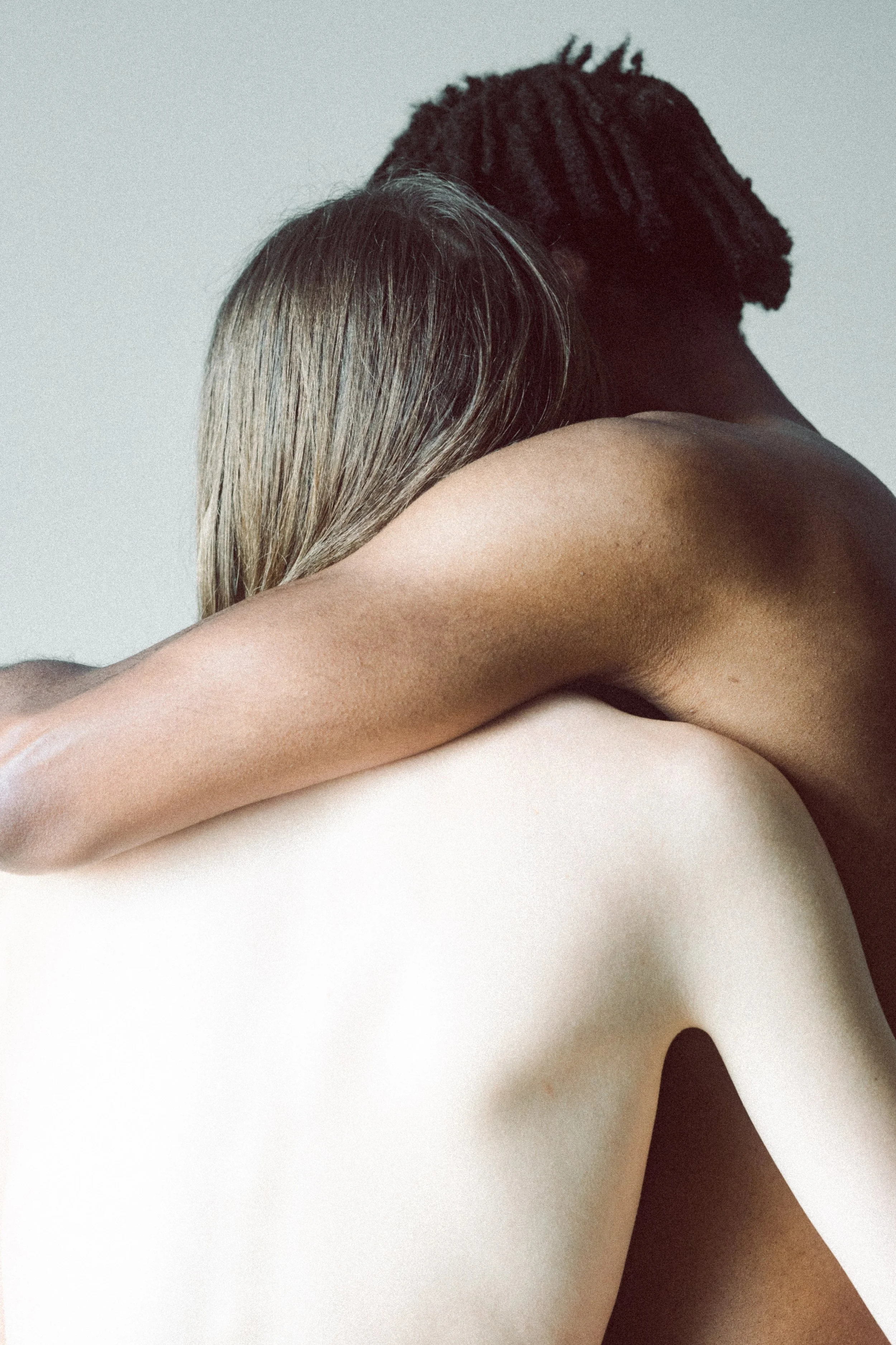 Close-up of two people embracing, with one person's head resting on the other's shoulder, showing their arms and partial back, against a plain background.