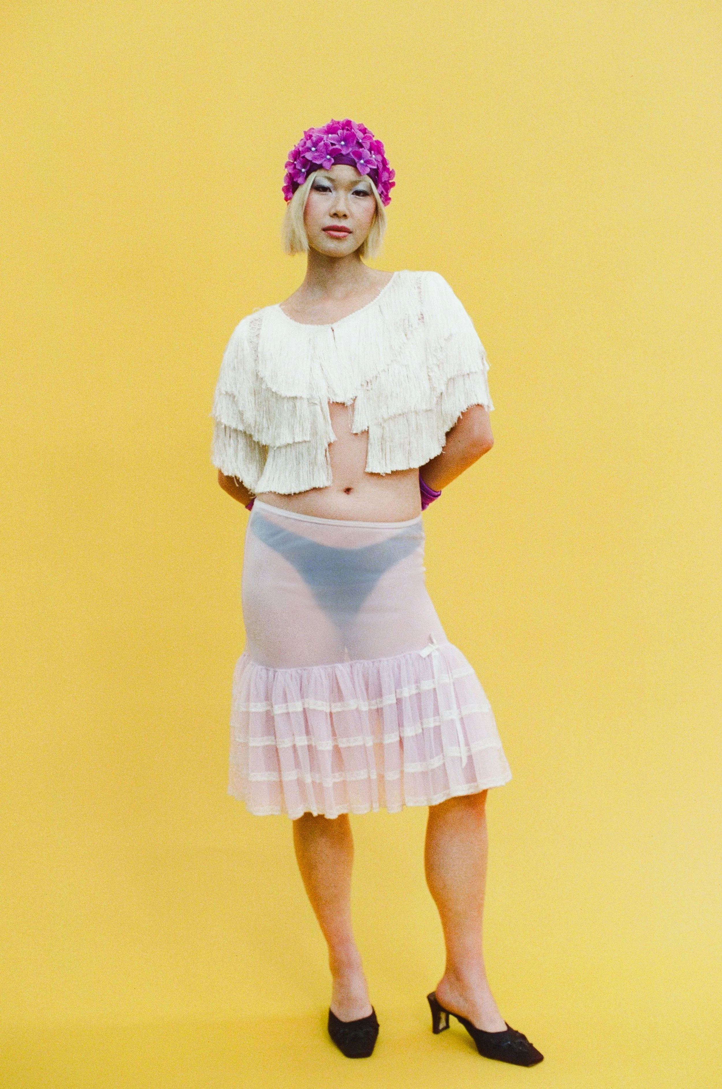 Editorial fashion portrait of a model wearing a white cropped blouse and sheer layered skirt, photographed against a vibrant yellow studio background.